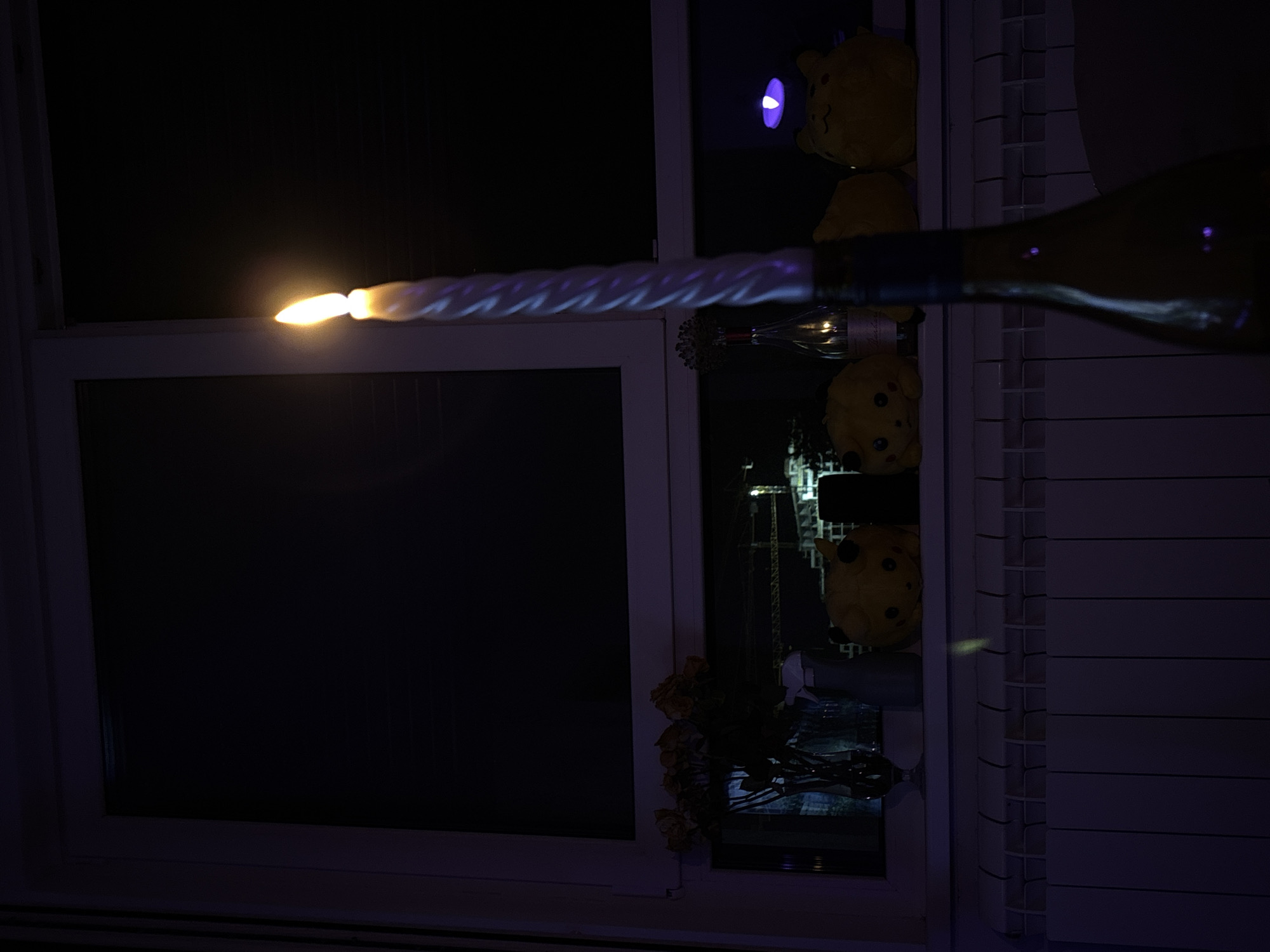 A lit white spiral candle stands vertically in a dark room, positioned in front of a window. The candle's flame is bright and casts a glow. Behind the candle, on a windowsill, are several objects. To the left, a vase holds a bouquet of wilted yellow roses. Next to the roses, a spray bottle is visible. Further to the right, two yellow stuffed toys resembling Pikachu sit side-by-side. Between the stuffed toys, a glass bottle with a label is placed, and a faint light emanates from within it. A purple light source is also visible on the windowsill to the right of the Pikachu toys. Outside the window, at night, construction cranes and building lights are discernible. In the foreground, a white radiator spans the width of the lower part of the image. A wine bottle is partially visible in the foreground, obscuring the radiator in places. The overall scene suggests an indoor setting at night, likely an apartment in the Zvezdara Urban Municipality, Serbia, given the characteristic radiator design and the view of urban construction.