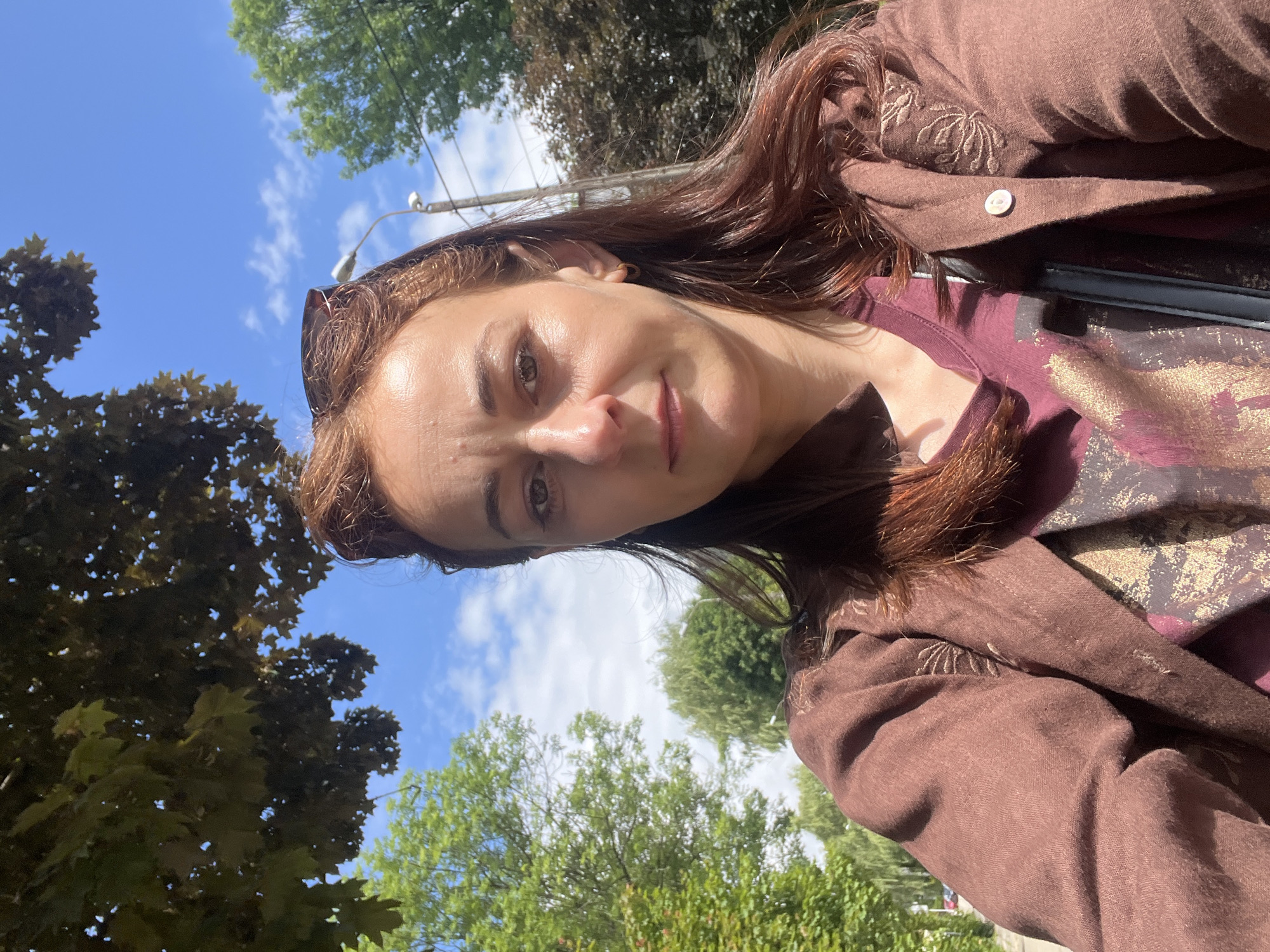 A close-up selfie captured outdoors on a bright, sunny day shows a woman looking towards the camera with a subtle, gentle smile. Her head and upper torso are visible, positioned slightly to the left side of the frame. She has medium-length brown hair, pulled back from her face, with sunglasses perched on top of her head. Her eyes are open, and her face shows natural skin tones with some shine, indicative of sunlight.
She is wearing a brown, possibly linen, button-down jacket or shirt, featuring light-colored embroidery of what appears to be a palm frond motif on the upper arm/shoulder. Beneath the jacket, she wears a burgundy or maroon t-shirt with a shimmering gold or bronze pattern. A single white button is visible on her outer garment.
The background features a vibrant blue sky dotted with scattered white clouds. Lush green trees frame the scene, suggesting an outdoor environment like a park or a tree-lined street. A utility pole with wires and a street light fixture is visible against the sky in the upper central part of the image. There is no visible text.