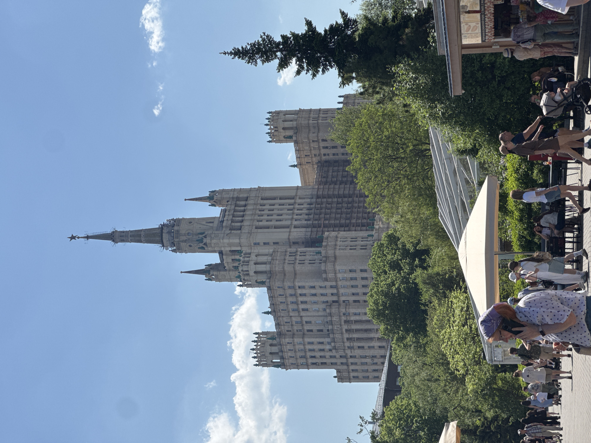The image showcases a street scene in Moscow, Russia, featuring one of the Seven Sisters Stalinist skyscrapers, likely the Hotel Ukraina, in the background. The weather is sunny with a blue sky and scattered clouds.
The foreground depicts a bustling area with many pedestrians. People are walking along a paved pathway lined with trees, adding greenery to the scene. A tent or structure with a white awning stands alongside the path, suggesting a market or event. Individuals are dressed in casual attire. The image captures a mix of locals and tourists.