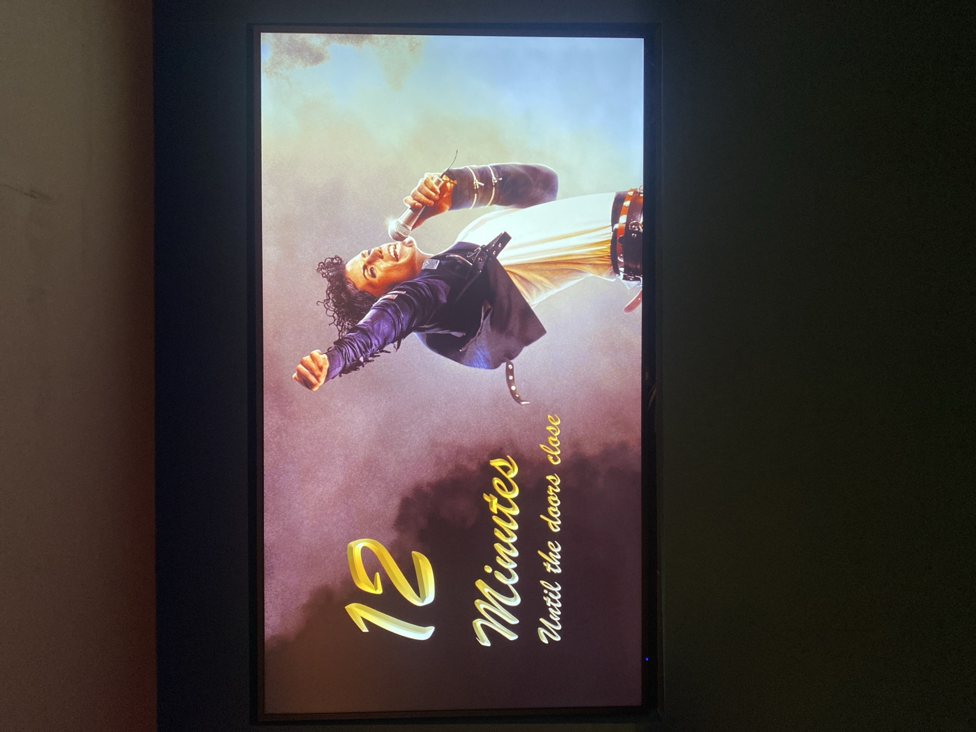 The media file presents a portrait-oriented image displayed on a dark-framed screen, captured in London, United Kingdom. The main subject on the screen is an artistic rendition of a person strongly resembling Michael Jackson, depicted mid-performance. He has curly dark hair, is wearing a dark jacket over a white, flowing garment, and appears to be in a dynamic, leaning-forward pose with one arm outstretched. His left hand holds a silver microphone near his open mouth, suggesting he is singing with passion, eyes closed.
The background of the image transitions from a bright, light blue sky at the top to a dramatic, smoky reddish-brown or purple hue at the bottom, contributing to an epic or ethereal atmosphere. The visual style is illustrative, resembling a digital painting rather than a photograph.
Prominently featured on the bottom right of the screen, overlaid on the background, is a countdown message in stylized, golden script. It reads "12 Minutes Until the doors close". This text indicates an imminent event, suggesting the display is part of a timed entrance or show.
The screen is bordered by a black frame. To the left of the screen, a plain, light brown wall is visible. The area to the right of the screen is dark. A small, faint blue light is visible at the bottom right corner of the screen's frame, indicating that the display is powered on.