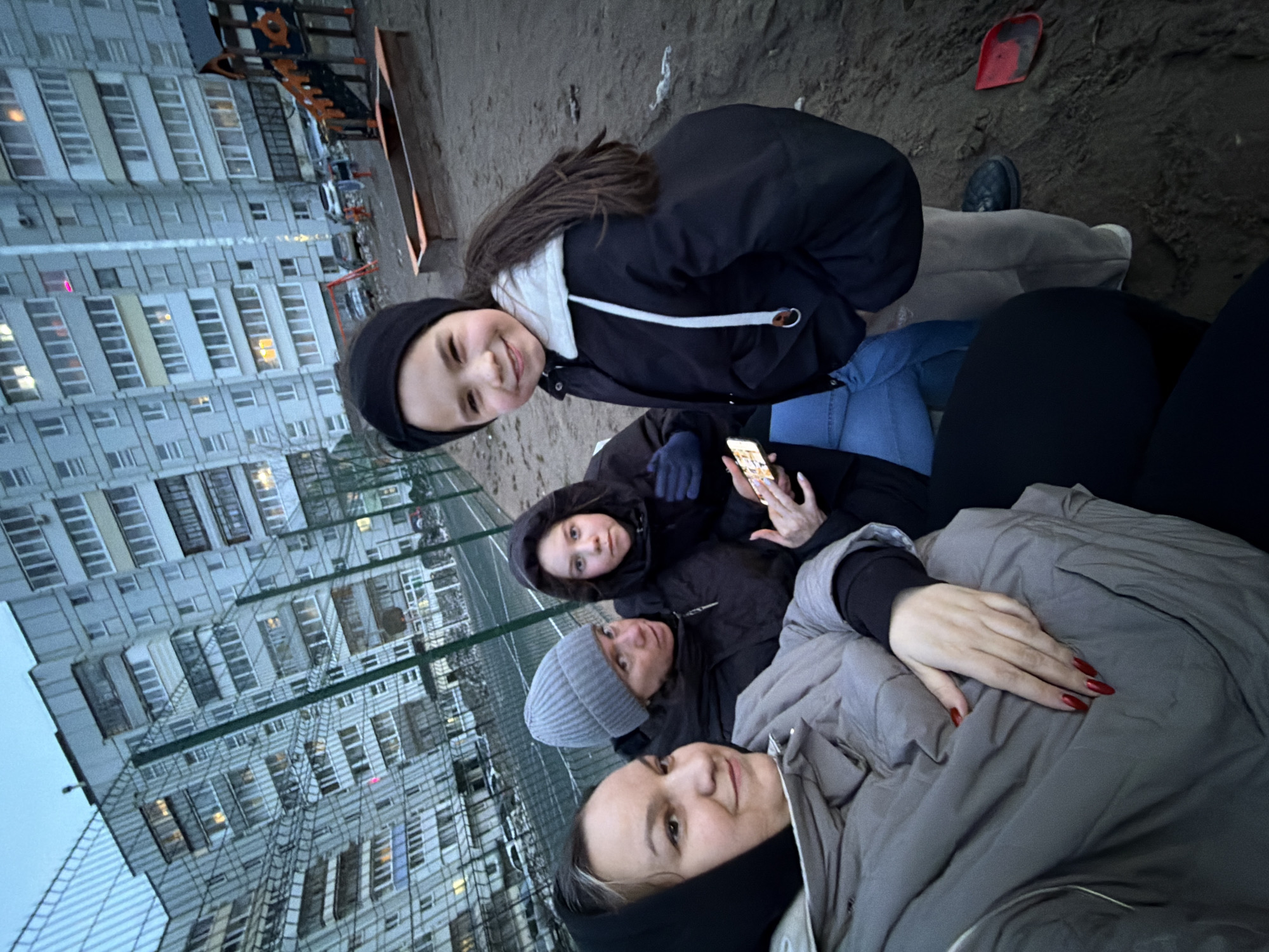 A group selfie or close-up shot, tilted on its side, features four individuals—three women and one young girl—outdoors, possibly in Chaikovsky, Russia. The setting appears to be a courtyard or common area beside large, multi-story apartment buildings, characteristic of Soviet-era architecture, with a green mesh fence separating the subjects from the background structures.
In the foreground, one woman with a black hoodie and a grey puffer jacket looks at the camera with a subtle smile. Her hand, with red manicured nails, rests on her jacket. Next to her, another woman in a grey beanie and black jacket looks upwards. Behind them, a third woman, dressed in a black hoodie and blue gloves, is focused on a smartphone held in her hands. The young girl, positioned to the left (when the image is upright), wears a black beanie, a black jacket with a white hood, and blue jeans, smiling widely at the camera.
The time of day appears to be dusk or early evening, indicated by the dim, overcast light and illuminated windows in the distant apartment buildings. The subjects' warm clothing (jackets, beanies, hoodies, gloves) suggests cool weather. The ground is a dark, sandy or dirt surface, and a red shovel is visible near the top right of the frame. Part of a playground structure can be seen behind the fence. No visible text is present in the image.