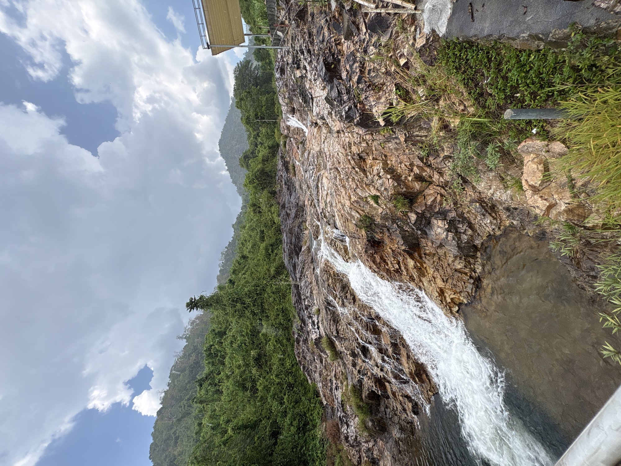 The image depicts a waterfall in Khanh Vinh, Vietnam. Water cascades over a rock face, bordered by lush green vegetation. The sky is overcast with patches of blue. A man-made structure is visible at the top left of the frame. The water collects in a pool at the base of the falls.
