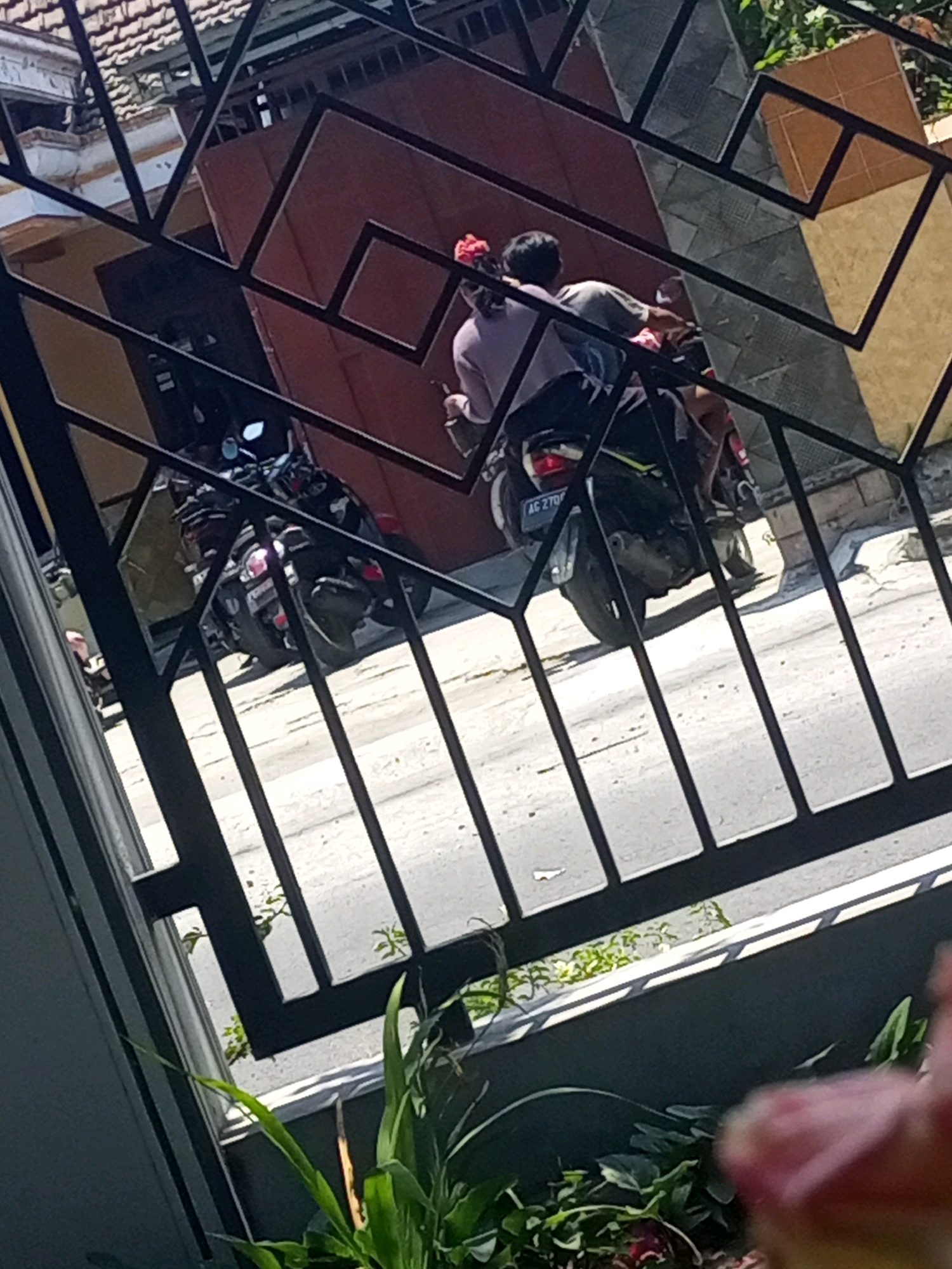 In Ngasem, Indonesia, two individuals are seated on a parked motorcycle, viewed through a metal fence. The person in front wears a light purple shirt, and the person behind has a red accessory in their hair. The motorcycle is dark-colored with a yellow accent and has license plate "AC 270F". Behind them, a large, dark red door is visible. The scene is outdoors, with sunlight casting shadows. Green foliage is present in the foreground and background.
