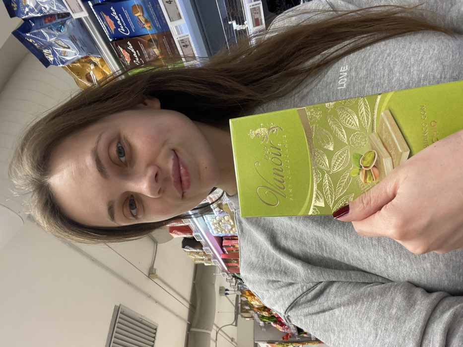 A female individual is depicted in a supermarket in Monsummano Terme, Italy, holding a bright green box of "Vanoir FINE CHOCOLATE" with gold accents. The box front displays illustrations of cocoa pods and two white chocolate bars with pistachios, bearing the partial text "BIANCO CON PISTACHIO". She wears a grey sweatshirt with "LOVE" embroidered on the left chest and has dark red nail polish on her left hand, which holds the chocolate. Her long brown hair frames a face with light-colored eyes, and she exhibits a slight smile directed towards the viewer. The background reveals typical supermarket shelving stocked with various packaged food items, including other chocolate brands like Venchi and Quadro, with visible price tags such as €4.95 and €2.99. Overhead fluorescent lighting and a ceiling ventilation grille are also visible within the store environment.