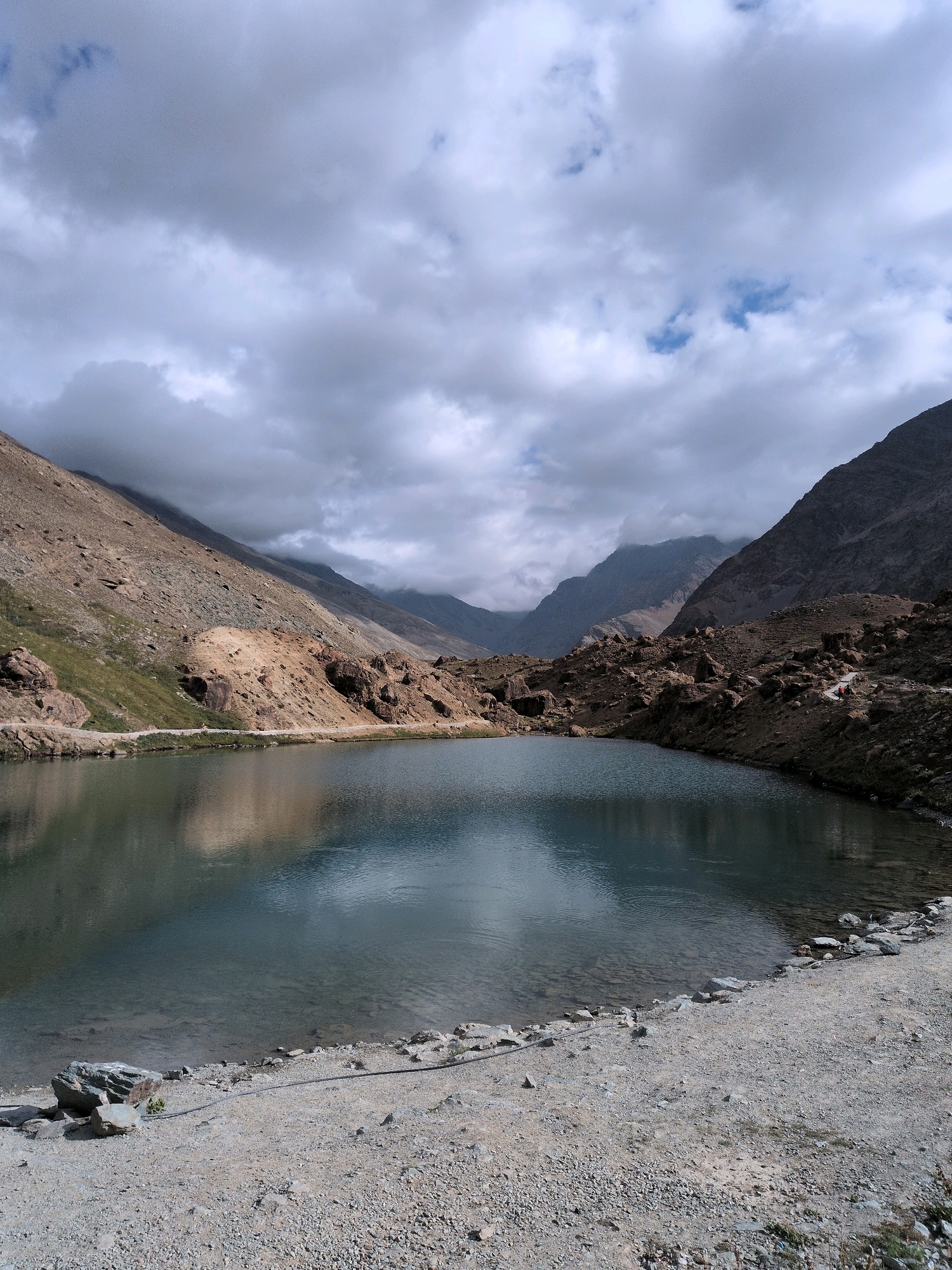 This image depicts a serene mountain lake in Patseo, India, under a dramatic sky. The lake, with its clear, reflective waters, occupies the foreground, bordered by a gravelly shore dotted with rocks and some sparse vegetation. In the midground, rugged, rocky hills rise steeply on either side of the lake. The left side features a mix of scree and patches of green grass, while the right side is characterized by a more barren, rocky slope. In the distance, a chain of majestic mountains stretches across the horizon, shrouded in a layer of cloudy mist, adding to the atmospheric depth of the scene. The overall impression is one of raw, natural beauty and tranquility.