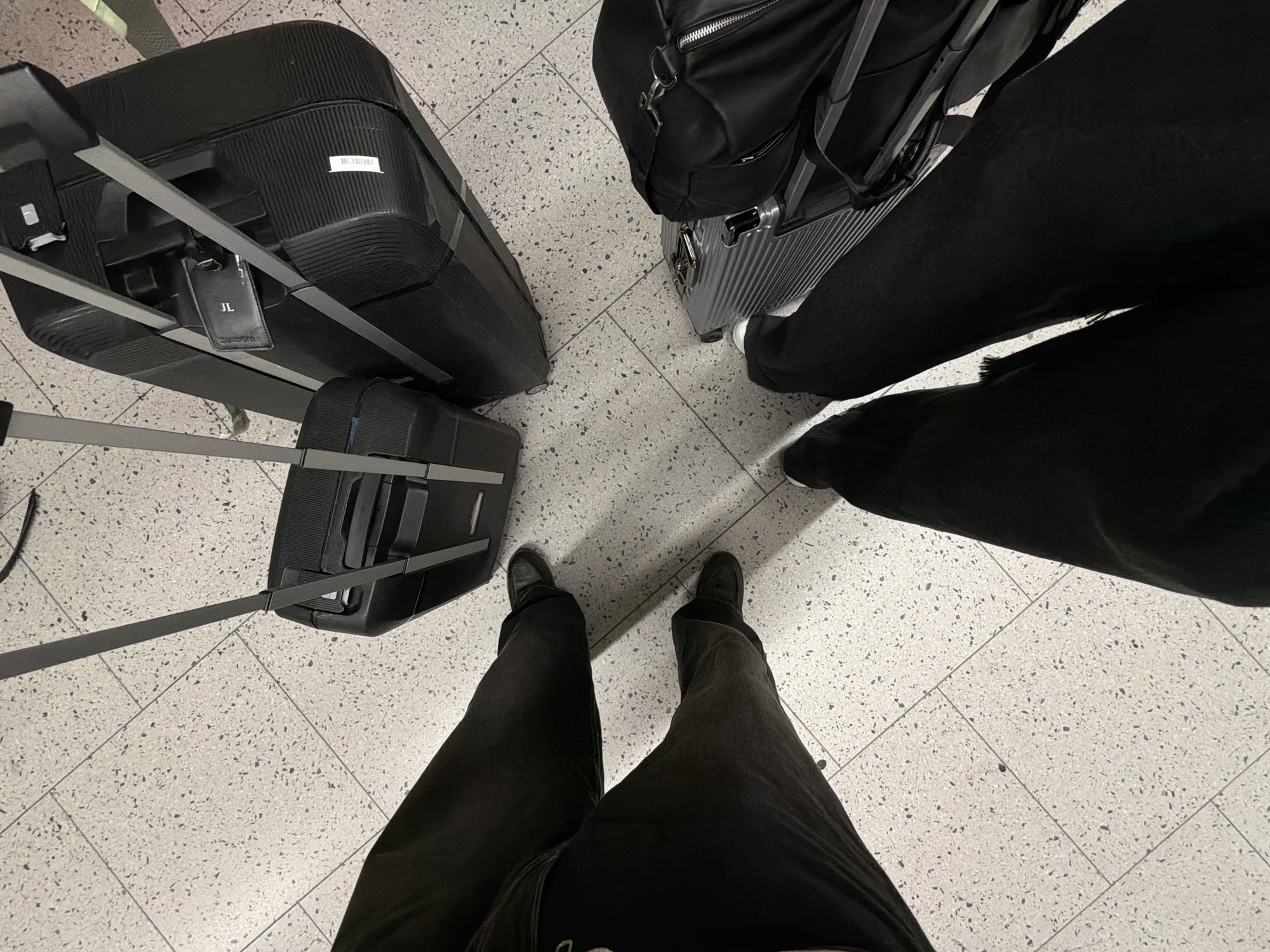 The image depicts two individuals standing on a light-colored, speckled tiled floor, likely within an airport terminal in Crawley, United Kingdom, given the presence of luggage. Both individuals are wearing dark clothing. One person's legs and feet, clad in dark pants and black shoes, are visible in the foreground on the left. Their feet are positioned on the tiled floor. In the mid-ground, a large black suitcase with a retractable handle and a small black luggage tag with a 'JL' initial is situated. Further back, another black suitcase is visible. On the right side of the image, the legs and feet of the second individual are seen, also wearing dark pants and white-soled shoes. They are standing next to a silver-colored wheeled suitcase with a black bag placed on top. The scene suggests a travel context, possibly at Gatwick Airport.