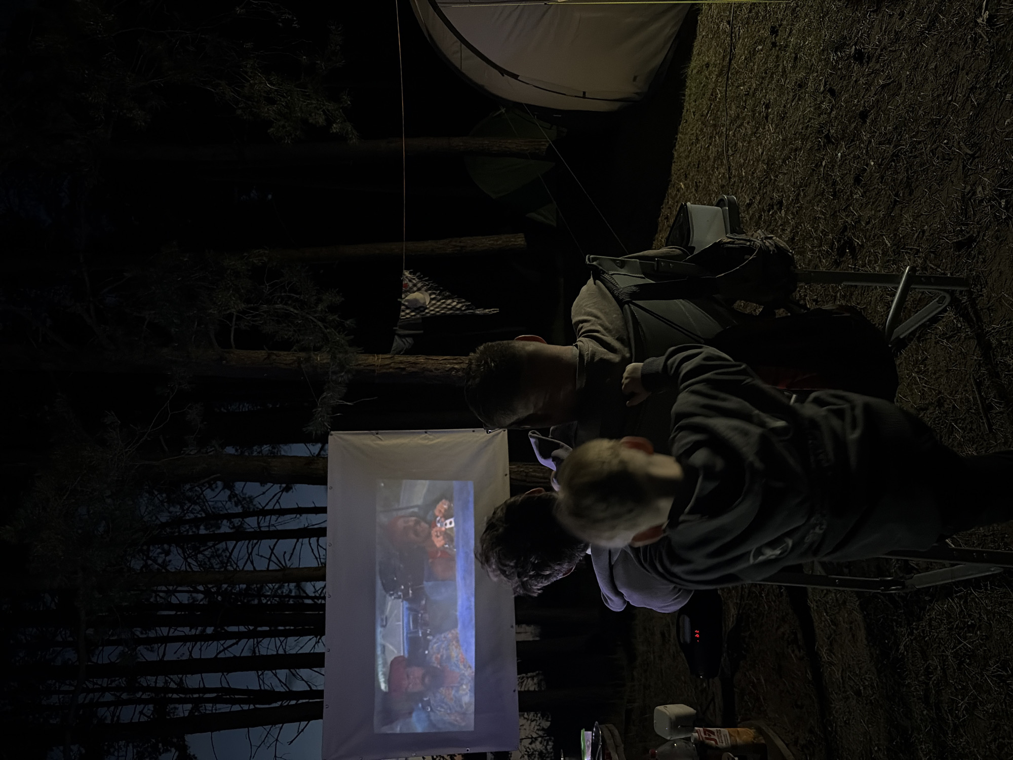 Three people are gathered in a dimly lit outdoor setting, possibly a campground or park, watching a movie projected onto a white screen. The projection shows an indoor car scene, with two actors in it. Two tents are visible in the background, one of which is green. The people are positioned near the screen, with one facing forward and the other two leaning in to view the screen. The environment includes trees, grass, and camping equipment, suggesting they are outdoors at night. Based on the request, the location is Zabegaevo, Russia.