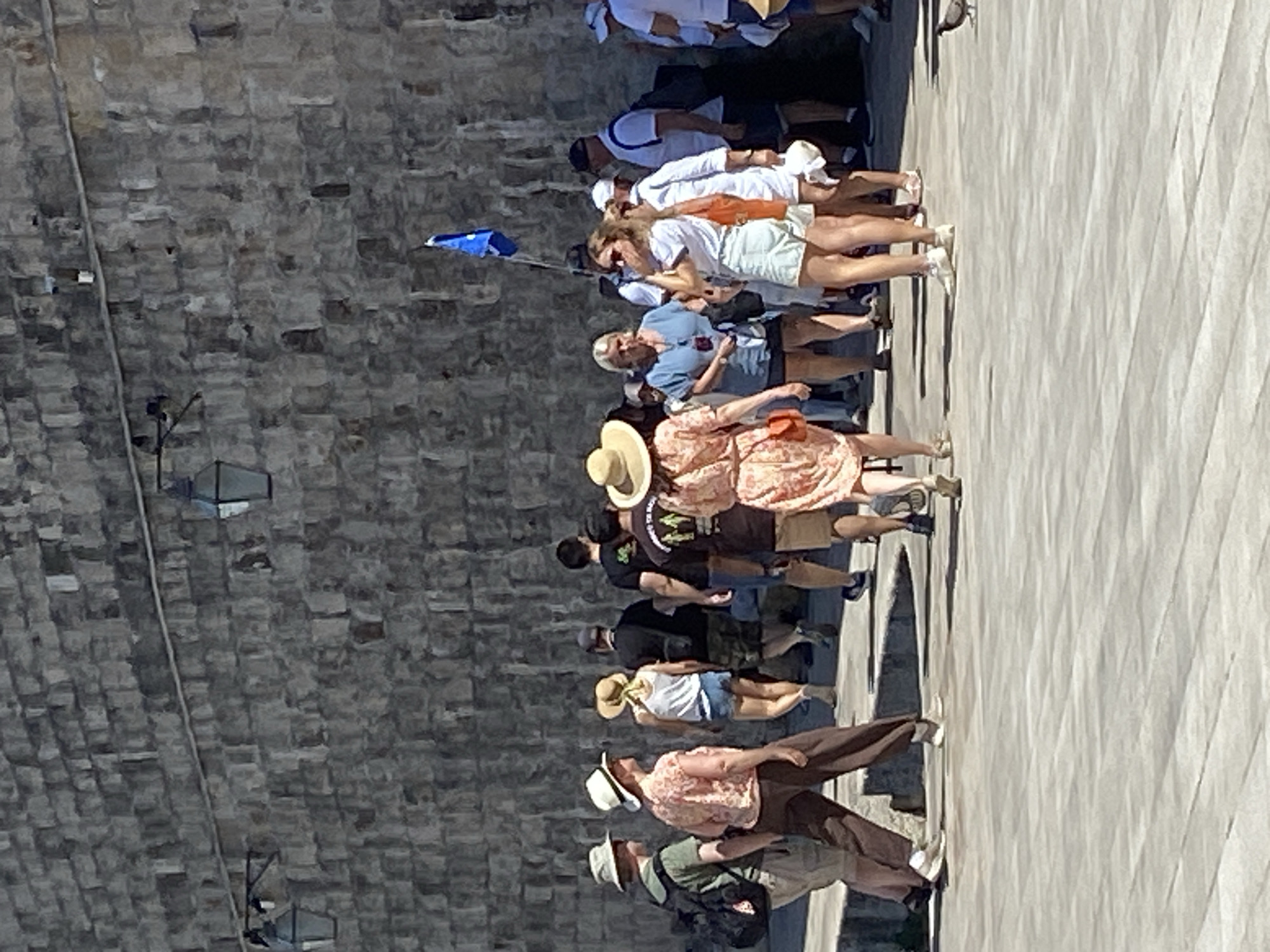 The image shows a group of tourists walking along the stone walls of Dubrovnik, Croatia, on a sunny day. The wall, constructed of roughly hewn grey stone blocks, dominates the background. Two dark, wrought-iron lanterns are mounted on the wall. In the mid-ground, a diverse group of people are seen from the waist down, walking on a paved stone area. They are dressed in casual summer clothing, including shorts, skirts, and t-shirts, and several are wearing hats, suggesting protection from the sun. A bright blue flag on a pole is visible in the upper right portion of the image, held by an unseen person. The ground is paved with large, light grey rectangular stones. A single pigeon is visible on the ground in the lower right corner.