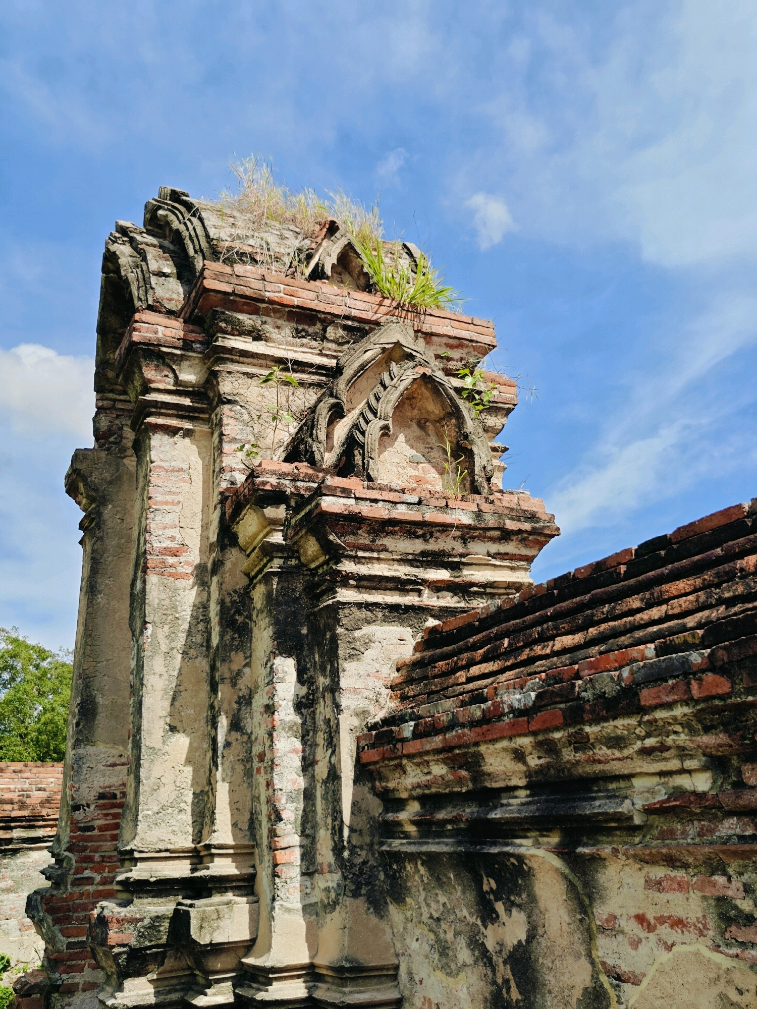 The image shows a close-up, low-angle view of an ancient brick structure, likely part of a temple ruin, under a partly cloudy blue sky. The structure is made of weathered red bricks and has decorative elements, including arched indentations and layered ledges. Vegetation, including grass and small plants, is growing from cracks and on top of the structure, indicating age and disrepair. The bricks are aged and discolored, with some areas showing moss or lichen growth. In the background, glimpses of more brickwork and green foliage are visible, suggesting a larger complex or ruins. The scene conveys a sense of historical decay and the enduring presence of nature reclaiming man-made structures. This image is likely from Ayothaya Town Municipality, Thailand, given the architectural style commonly found in the ancient ruins of this region.