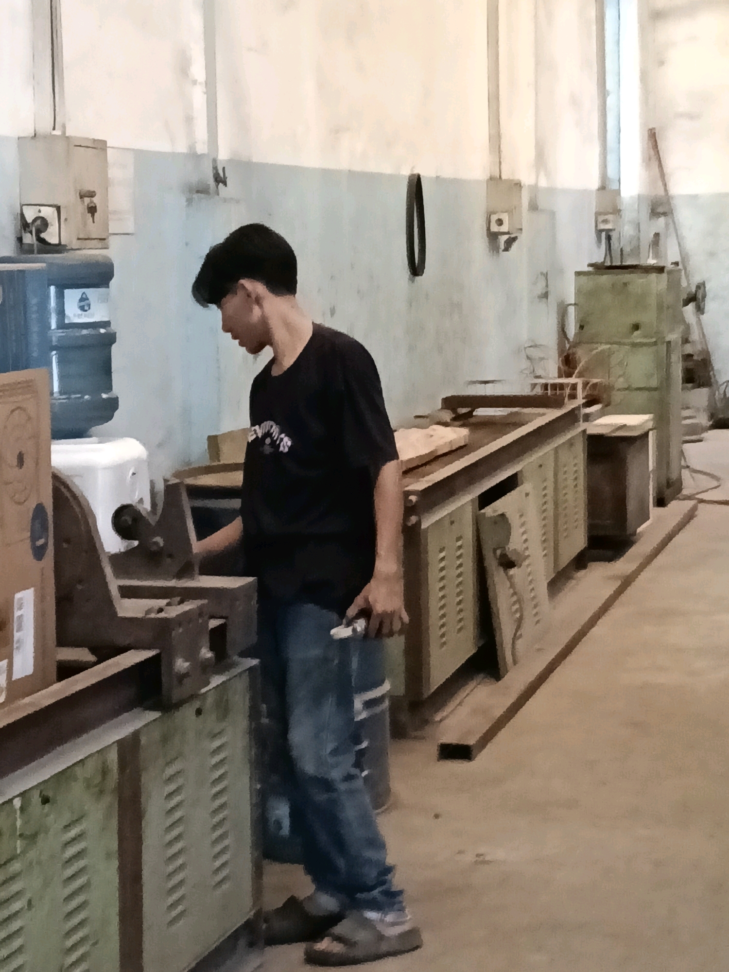 A young male, appearing to be of Southeast Asian descent, stands in what looks like a workshop in Belimbing, Indonesia. He is wearing a black t-shirt and blue jeans. The male is seen interacting with a piece of machinery, holding a tool in his right hand. The environment appears to be a functional space with various equipment, including what seems to be a water dispenser with large blue containers, several workbenches, and metal cabinets. The lighting suggests an indoor setting, possibly a factory or a large workshop. The walls are painted a pale blue and white, with some electrical boxes mounted on them. The floor is concrete.