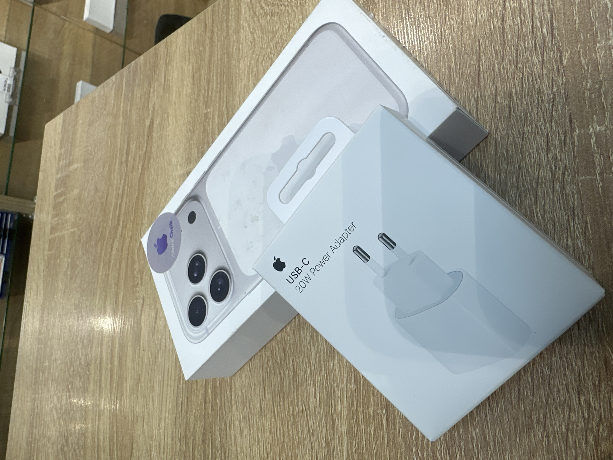 The image depicts two Apple product boxes on a wooden surface. One is a clear iPhone case with a sticker from "iStore Chaikovsky." The other box contains a USB-C 20W Power Adapter. The location, based on the "iStore Chaikovsky" sticker, is Chaikovsky, Russia. The scene includes no people or specific actions. The items are arranged on a wooden surface.