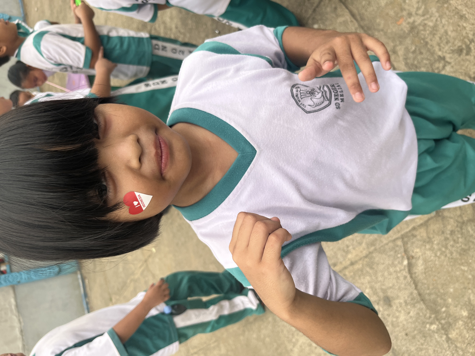 A young child with black hair and a sticker on their cheek that says "I love Indonesia" is looking at the camera. They are wearing a white shirt with green trim and green pants. The child is in what appears to be a schoolyard in Meliau Hulu, Indonesia, with other children in the background wearing similar uniforms. The ground is paved with concrete.