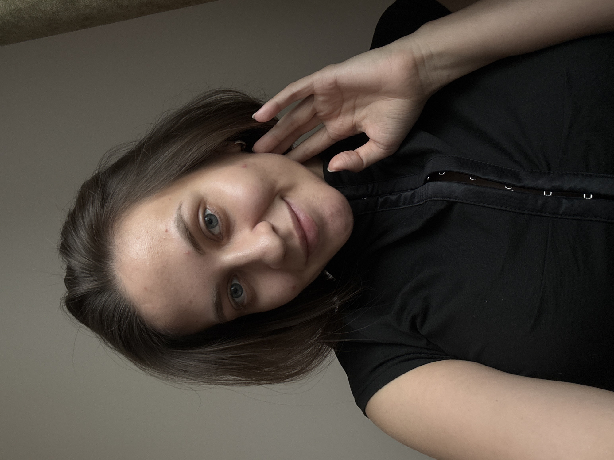 A young woman with short, brown hair and blue eyes is looking at the camera. She is wearing a black, short-sleeved shirt. Her right hand is raised to her face, touching her neck. The background is a plain, light brown wall with a curtain on the right side. The image appears to be a selfie taken indoors, with the woman's face and upper body filling most of the frame. There are no specific landmarks or environmental details that would indicate a location in the city of Pervomaysky, Russia.