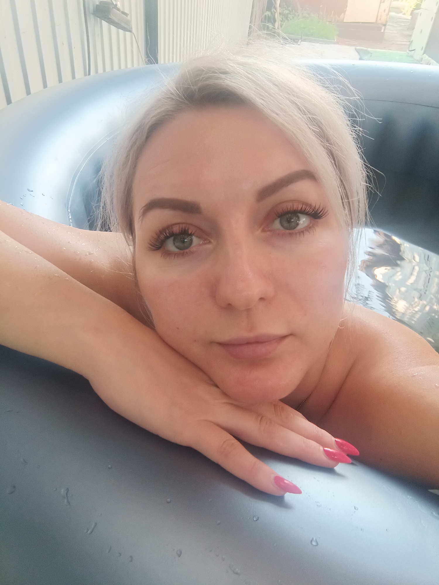 A close-up selfie of a blonde woman with green eyes and long, false eyelashes is taken from the chest up. She is leaning on the inflatable edge of a hot tub, with her arms crossed and her head resting on her hands. Her fingernails are painted bright pink. Water droplets are visible on the hot tub's surface and on her arms. The background shows a corrugated metal wall, suggesting an outdoor or semi-outdoor setting. The woman's hair is light blonde and slightly messy. The overall impression is casual and relaxed.