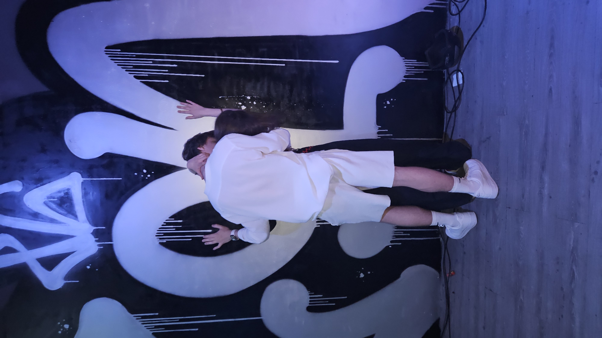 The image depicts a man in a white shirt, white shorts, white socks, and white sneakers lying on top of another individual on a black and white graffiti-covered surface. The graffiti includes large, rounded shapes and sharp, angular lines. The man is face down, with his hands spread on the graffiti. His head is positioned near the head of the person underneath him, whose dark hair is visible. The legs of the person underneath are clad in black. A pair of headphones and an amplifier lay to the side, alongside a grayish-blue floor. The location of the scene is unknown, and there is no way to tell that the location is Nha Trang, Vietnam.