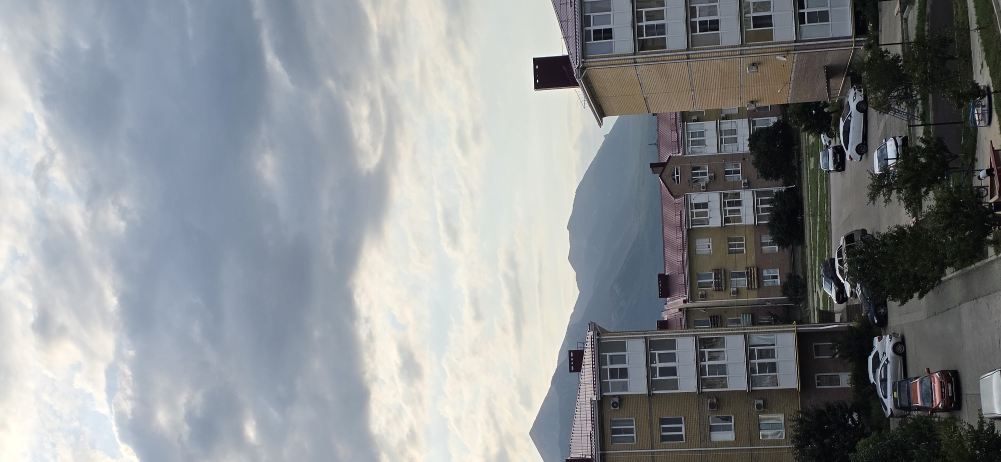 This is a vertical, medium-angle, outdoor, full shot of apartment buildings with a mountain range in the background. The sky is filled with heavy, gray clouds, with some patches of lighter sky peeking through. The apartment buildings are constructed with light yellow and beige bricks, and feature white-framed windows and balconies with glass enclosures. They have red tiled roofs. In the foreground and middle ground, several cars are parked on a paved area. These include a white sedan, a dark SUV, a silver sedan, and a dark gray hatchback. There are also trees and some playground equipment visible in the lower portion of the image. The mountains in the background are hazy and muted in color, suggesting distance and possibly an overcast day. The scene appears to be from a residential area in Inozemtsevo, Russia, likely taken in the late afternoon or early evening due to the lighting conditions.