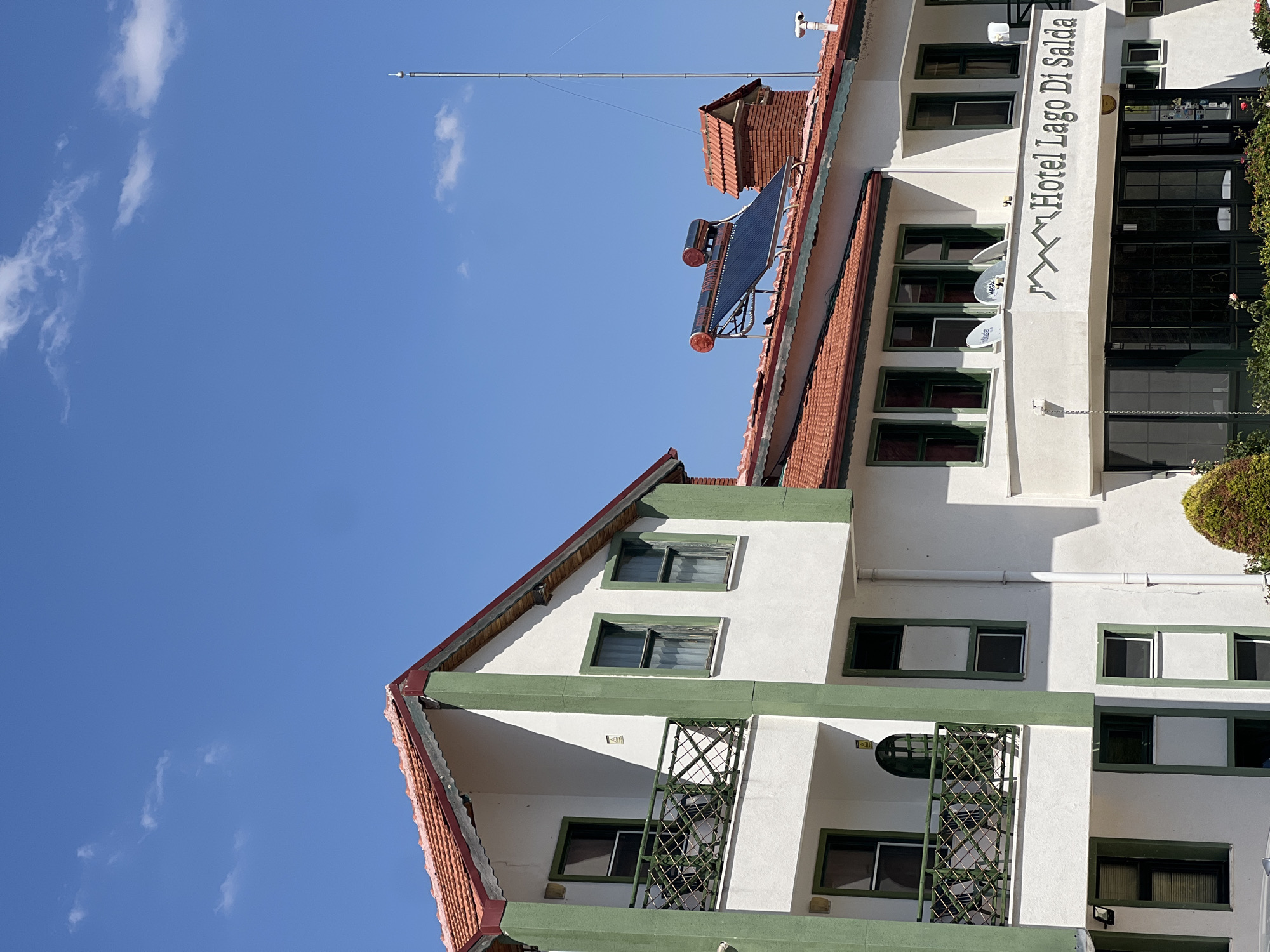 The image shows the exterior of the Hotel Lago Di Salda in Yeşilova, Turkey, under a clear blue sky with scattered white clouds. The hotel is a multi-story building with white walls and distinctive green trim around windows and vertical columns. The roof is covered in red tiles. On the roof, a solar water heater and a tall, slender antenna are visible. The hotel's name, "Hotel Lago Di Salda," is prominently displayed above the entrance. The ground level features large glass doors and windows, and a small, manicured bush is located in the foreground. The overall scene depicts a sunny day at a hotel.
