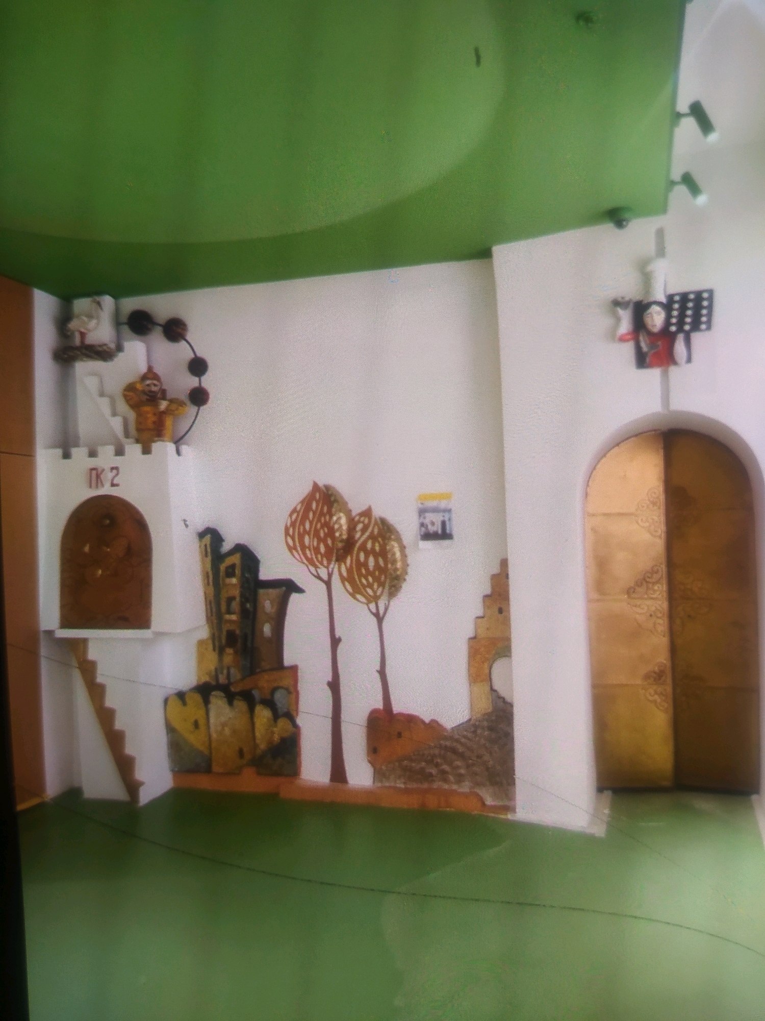 The image depicts a room with a green ceiling and walls decorated with various artistic elements. On the left wall, a white tower structure features an arched doorway with ornate carvings and the text "ПК 2" above it. A small, yellow-clad figure is perched on top of the tower. Next to the tower, a stylized representation of buildings is attached to the wall. To the right of these structures, two abstract, tree-like forms with patterned leaves are visible. In the center of the right wall, a large, arched, golden door is present. Above the door, a small decorative piece features a face and a grid-like pattern. The floor is a solid green. The scene suggests an indoor creative space, possibly a children's play area or an art installation, located in Birlik, Uzbekistan.