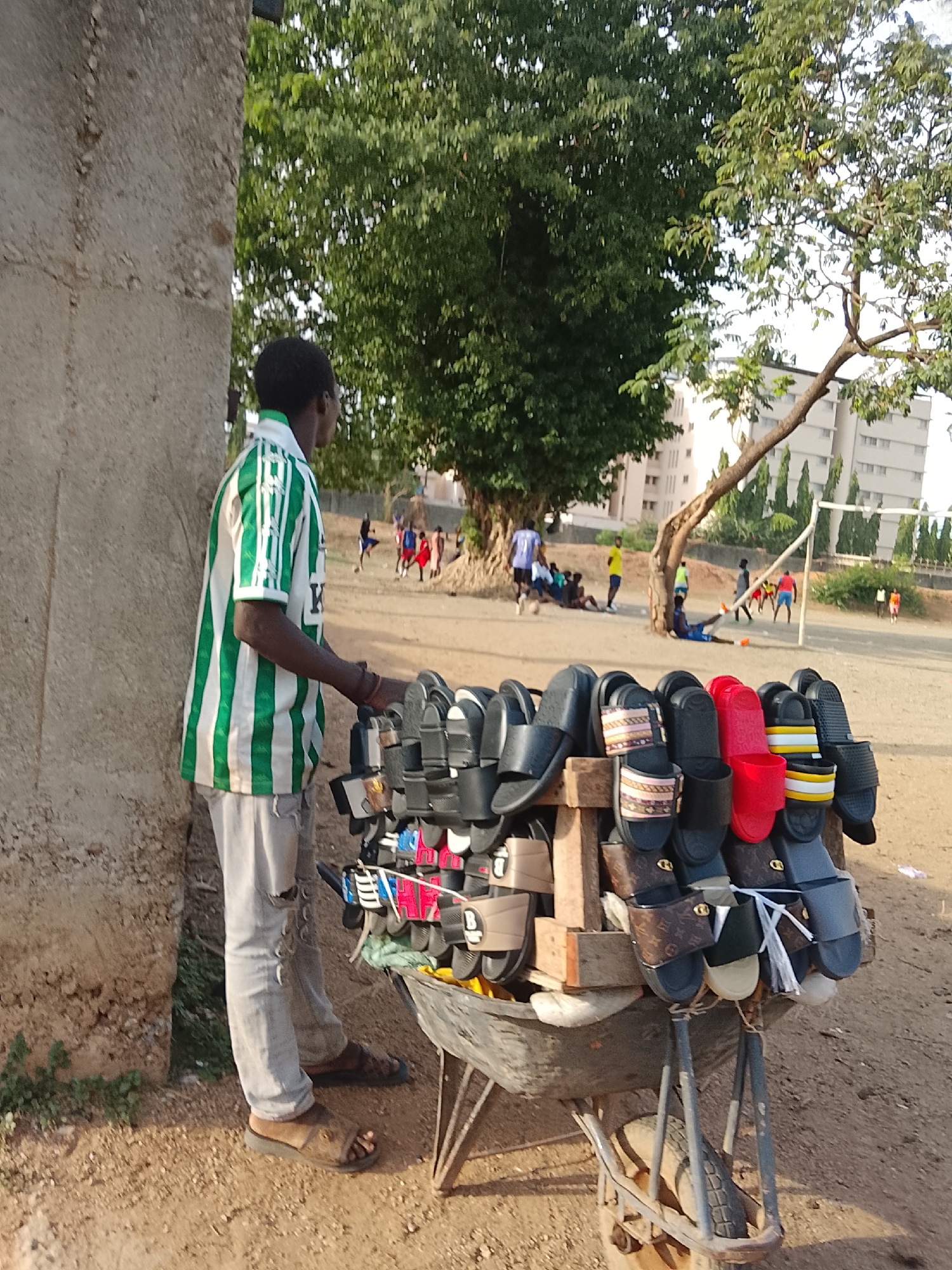 A street vendor wearing a green and white striped shirt and ripped light blue jeans stands near a concrete structure beside a dirt field in Abuja, Nigeria. He is next to a wheelbarrow loaded with pairs of sandals. In the background, a soccer game is underway, with multiple people playing and watching under the trees. A white building with multiple stories is visible in the distance, behind the soccer field and a soccer net with white posts. The ground is a mix of dirt and sand.