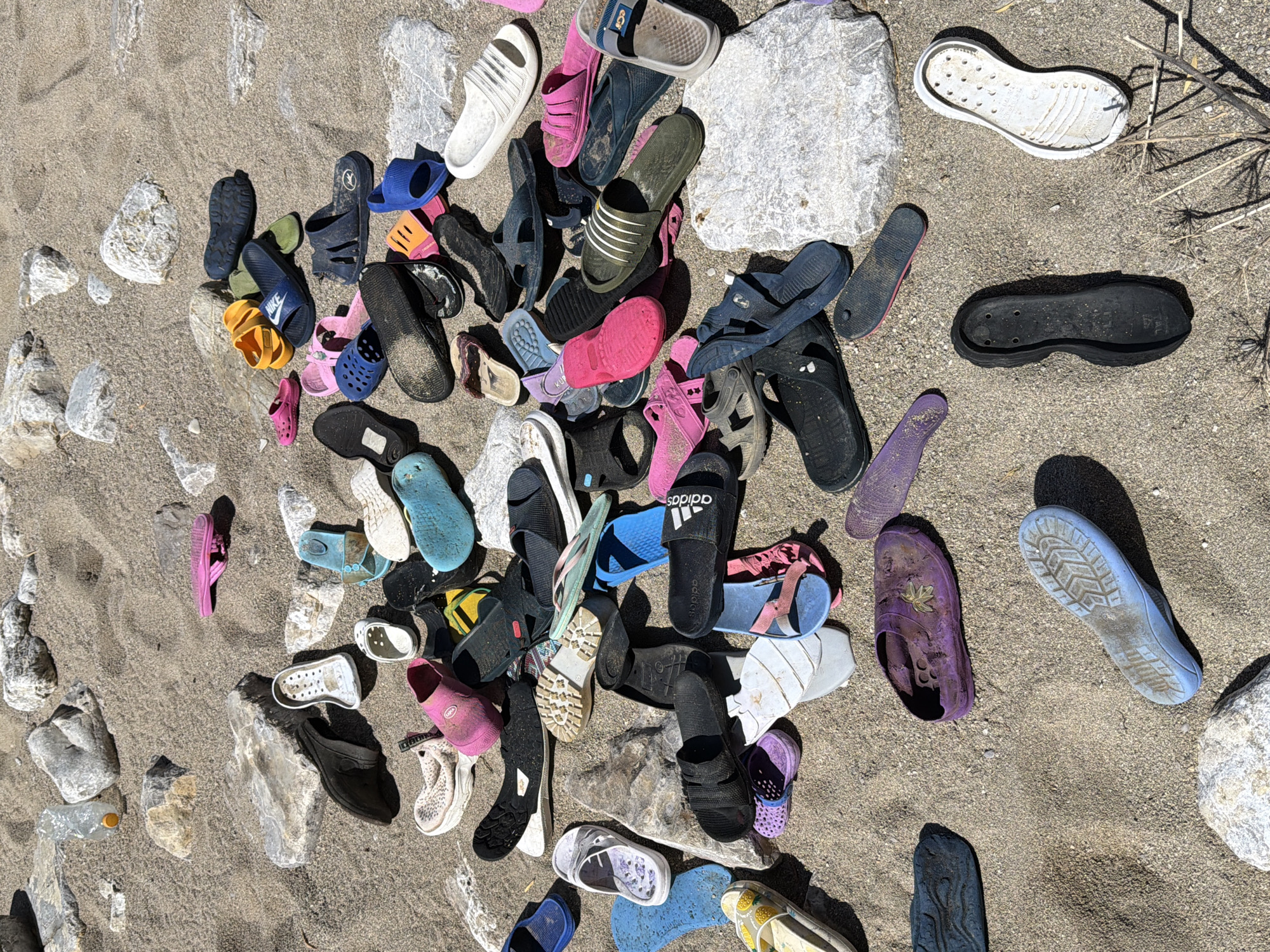 The image depicts a collection of discarded footwear scattered on a sandy surface, likely a beach. There are various types of footwear, including sandals, flip-flops, and Croc-style shoes, in different colors and sizes. The footwear appears worn and discarded. The sand is interspersed with pebbles and larger rocks. Based on the context, this scene is possibly located in Budva, Montenegro. No people are visible.