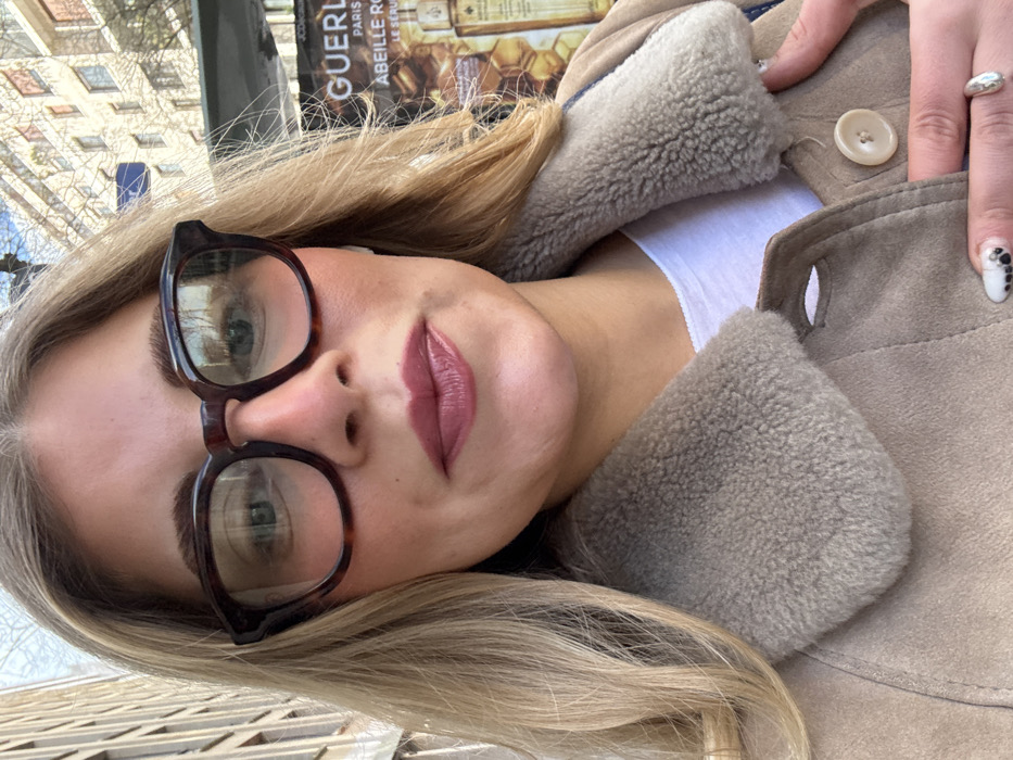 A close-up, eye-level portrait depicts an adult female positioned centrally. She has blonde hair, fair skin with visible blush, and wears square, tortoiseshell-patterned glasses. Her lips are coated with a glossy mauvy-pink lipstick. She is wearing a light beige or tan suede-like jacket featuring a prominent, plush, light grey sheepskin-style collar, over a white t-shirt. Her right hand, partially visible, rests on the jacket's lapel, revealing a white thumbnail with black irregular dots and a silver ring on her ring finger.
The background indicates an urban outdoor setting in Paris, France. To the right, a street advertisement is visible, prominently featuring "GUERLAIN PARIS" in black text, along with "ABEILLE ROYALE" and "LE SERUM," displayed over a honeycomb pattern with amber-toned product bottles. A small "JCDecaux" logo is also present on the advertisement. Above the advertisement and to the left, light-colored Parisian buildings with multiple windows and bare tree branches are visible against a bright sky, consistent with winter or early spring conditions.