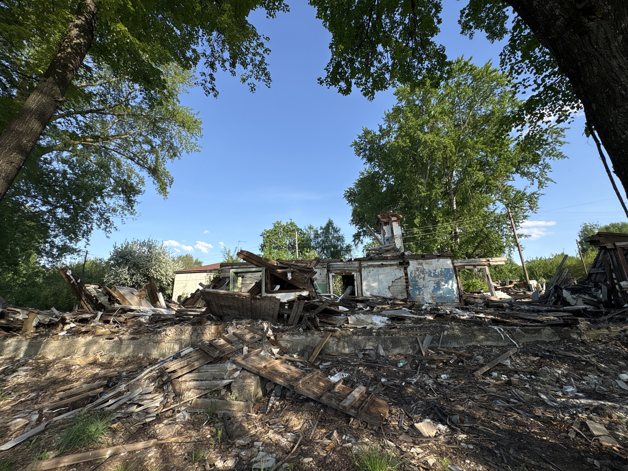 The image shows the ruins of a building, likely in Chaikovsky, Russia, given the prompt's instructions. The structure is largely demolished, with only sections of walls and the base of a chimney remaining. Debris, including wood planks and other building materials, is scattered across the foreground. The sky is visible through the destroyed structure, framed by trees. The scene suggests a recent demolition or a building destroyed by fire or natural disaster. The ground is littered with ash and fragments. There are no people visible in the image.