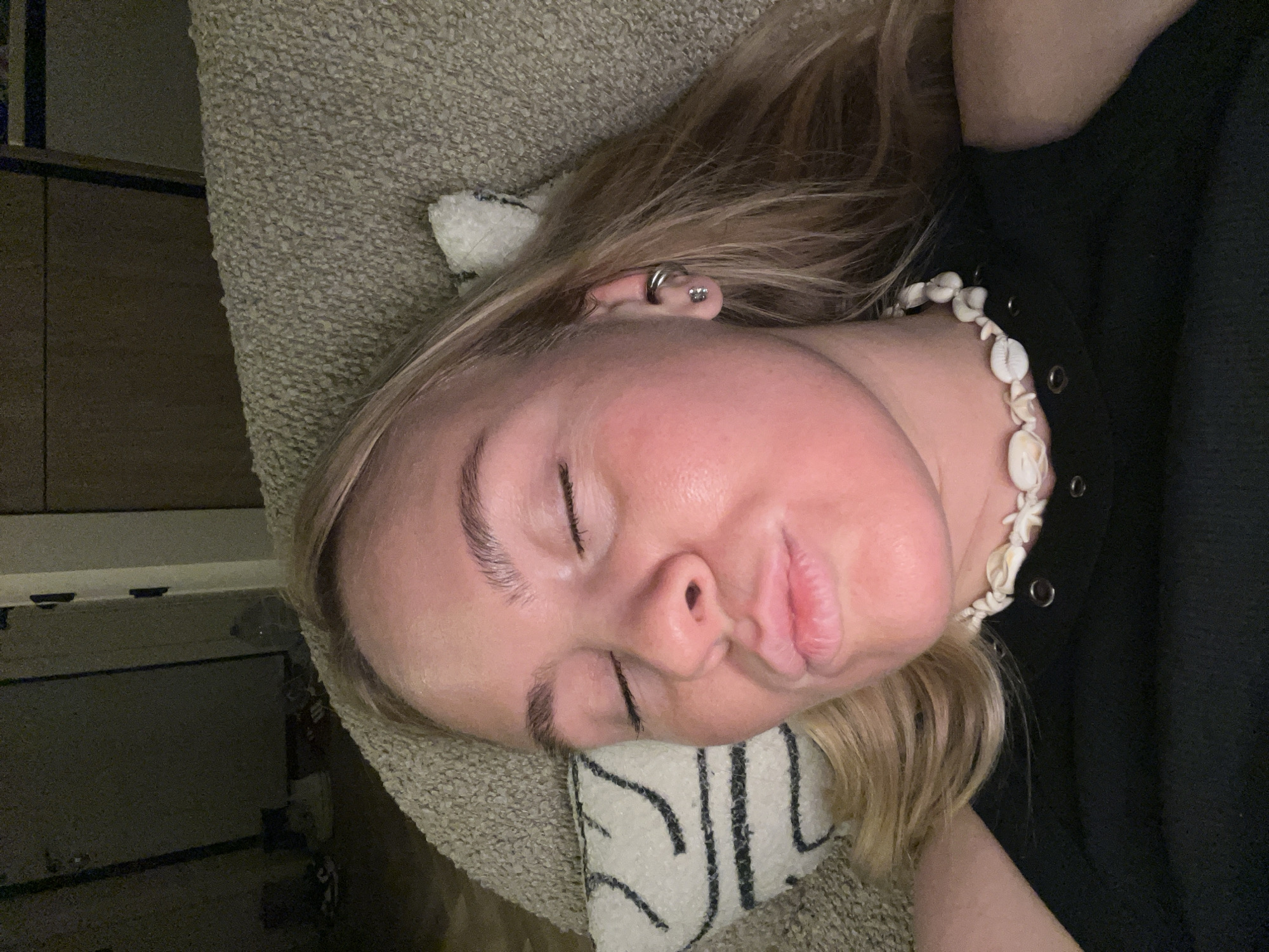 A woman is lying down on a textured surface. She has blonde hair and is wearing a black top with a white shell necklace. She has closed eyes.
There is no information available in the image to determine if the location is in the city of Рубцово, Russia.