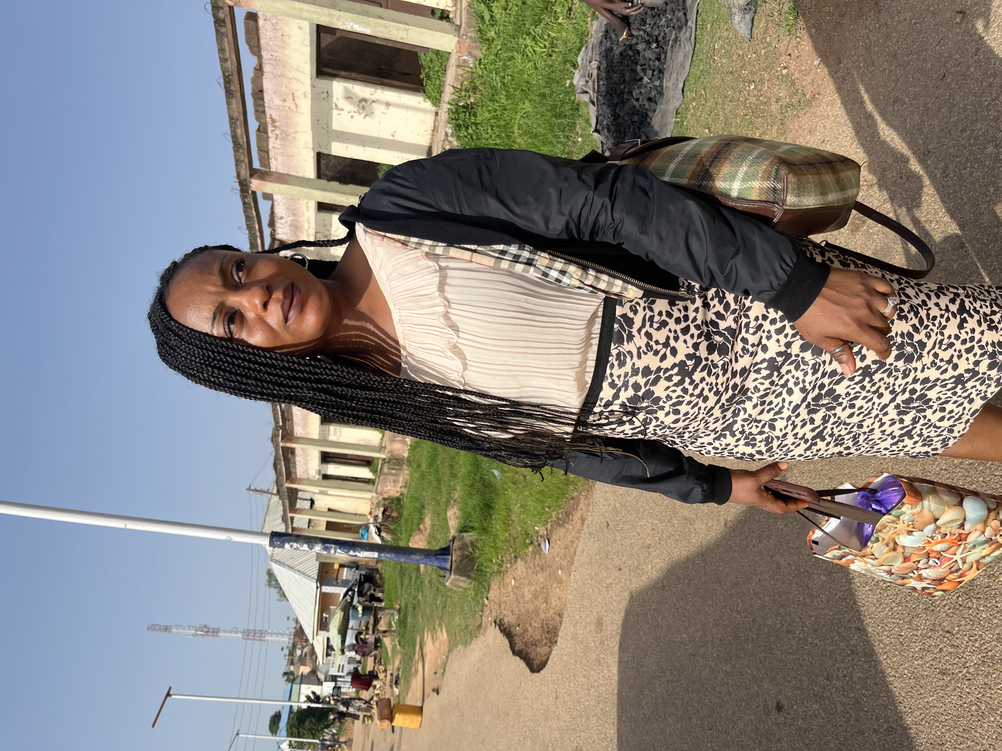 A woman with braided hair stands outdoors in what appears to be an urban setting in Jos, Nigeria. She is wearing a light-colored, ruffled blouse and a leopard-print skirt. A dark jacket is worn over her shoulders, and she carries a patterned shopping bag in her left hand and a plaid shoulder bag on her right.
The background shows a street with cracked pavement and sparse vegetation. Several buildings with weathered facades and open doorways are visible, suggesting some are under construction or in disrepair. In the distance, a cellular tower can be seen. Other people are present in the background, some near market stalls or engaged in unspecified activities. The sky is clear and blue, indicating a sunny day.