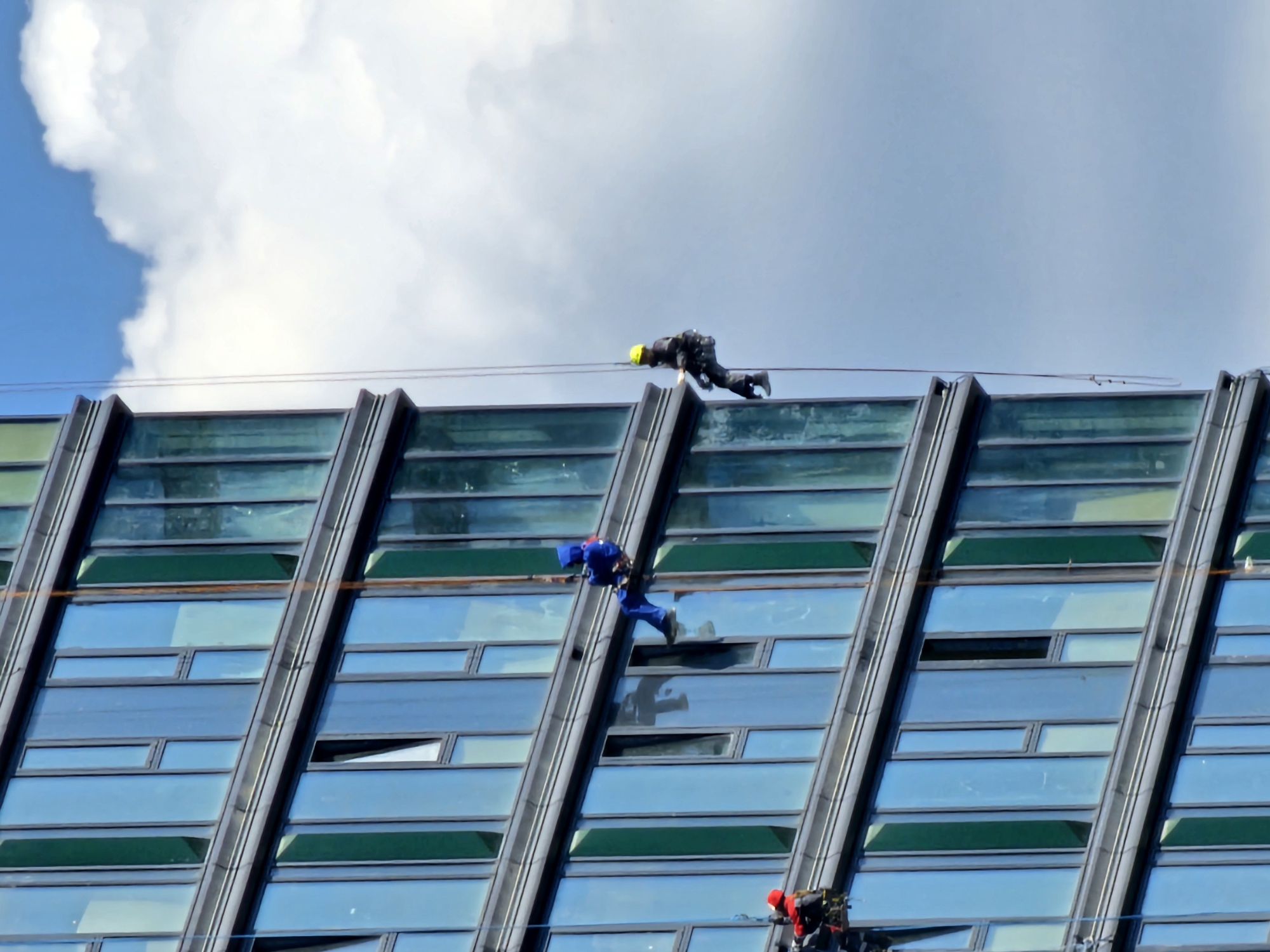 Three window cleaners are rappelling down the side of a modern, glass-faced skyscraper in Moscow, Russia. The building features numerous rectangular windows with a dark frame, set against a blue sky with scattered white clouds. The cleaners are suspended by ropes, wearing safety harnesses and work attire. One cleaner, wearing a blue jumpsuit, is positioned on the left side of the building, midway down. Another cleaner, in a yellow hard hat and dark work clothes, is rappelling to the right of the first, closer to the edge of the building. A third cleaner, wearing a red helmet and a dark suit with red accents, is visible further down and to the left, on a lower level of the building. The overall scene depicts a routine industrial task taking place against an urban backdrop.