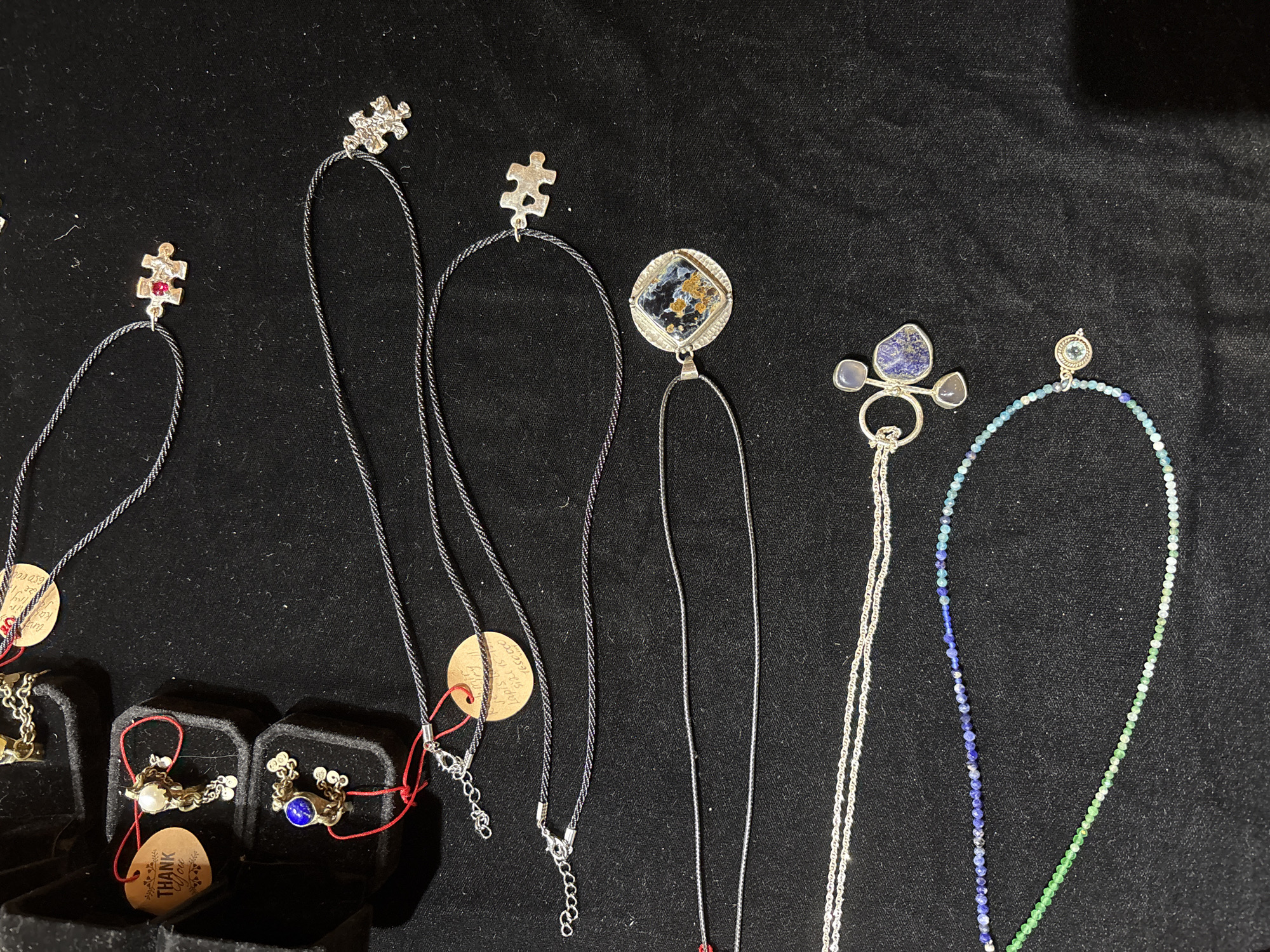The image shows a collection of necklaces displayed on a black velvet surface. The necklaces vary in design, with some featuring pendants and others made of beads. In the upper left corner, there are two small black velvet jewelry boxes, one containing a ring with a pearl and another with a ring featuring a blue stone. One box has a "THANK YOU" tag. There are also several tags with handwritten text scattered among the necklaces. The scene appears to be a display of jewelry, possibly at a market or a store. The location is given as Unknown, Unknown.