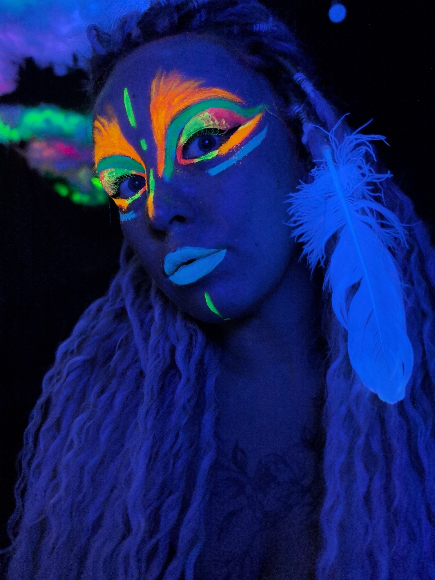 A person with blue-lit skin and light blue dreadlocks is centered in the frame against a dark background. The person's face is decorated with neon orange, green, and pink makeup, including a stylized design around the eyes and lips. A large blue-lit feather is visible near the right side of the face. The person is wearing an indiscernible blue-lit shirt. The location is not discernible and unknown.