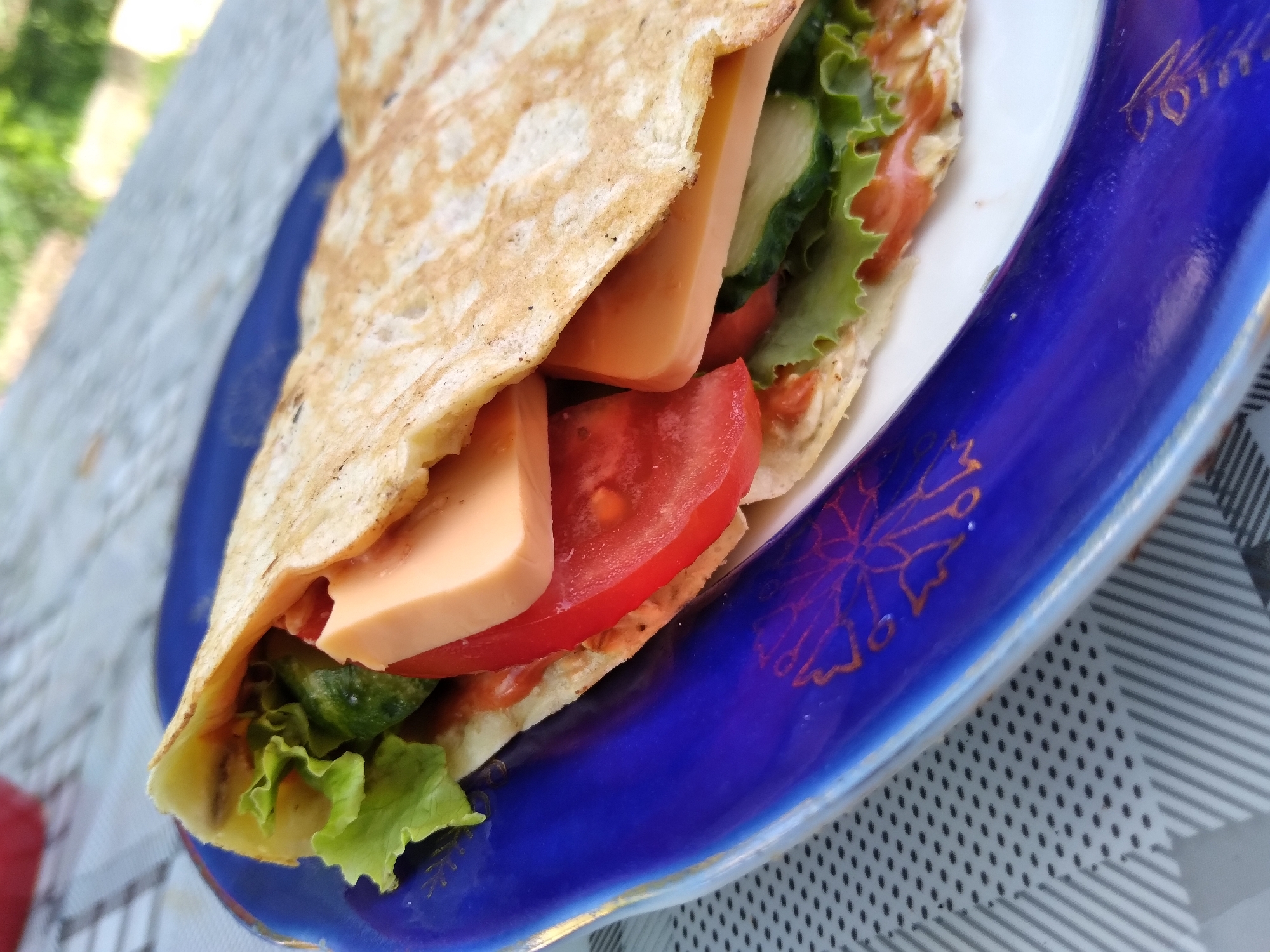 A close-up shot shows a savory crepe filled with lettuce, tomato, cucumber, and cheese. The crepe is folded in half and rests on a dark blue, oval-shaped plate with a gold rim and intricate floral designs. The plate is placed on a checkered tablecloth. The background is blurred, showing hints of green foliage and a hint of a red object on the left, suggesting an outdoor setting. The scene is set in Radisne, Ukraine.