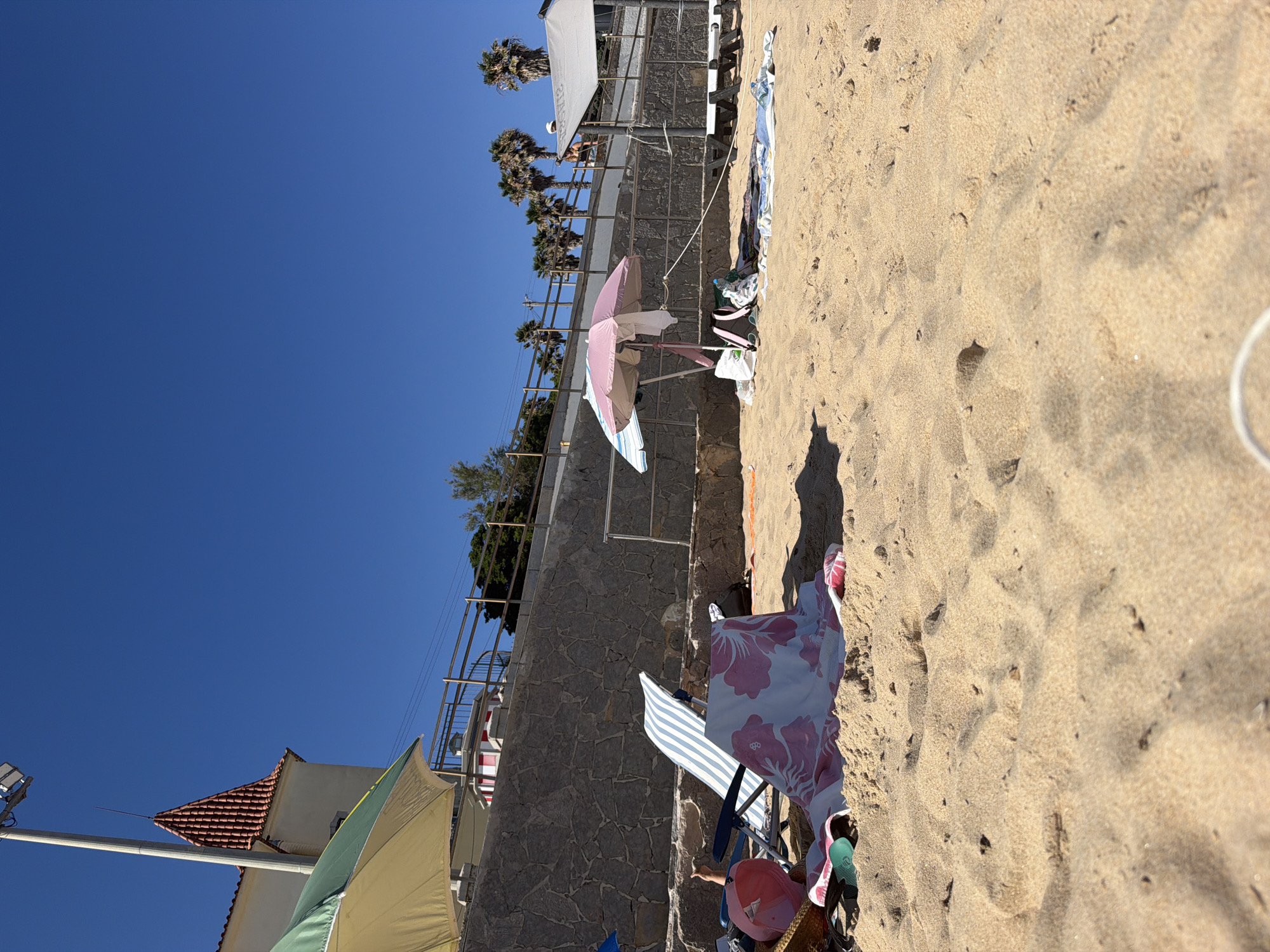 The image depicts a sunny day at a beach in Cascais, Portugal. The scene is oriented vertically. In the foreground, the sand is the primary subject, with footprints visible. A colorful beach umbrella, a striped beach chair, and various towels and personal items are scattered on the sand, suggesting a relaxed atmosphere.
In the middle ground, a stone wall runs along the coastline, separating the beach from a walkway or elevated area. Palm trees are visible on the elevated area, indicative of a warm climate. A pink and white beach umbrella is open, providing shade. Beachgoers can be seen in the distance on the elevated walkway.
In the background, a clear blue sky dominates the scene. A building with a red-tiled roof and white walls is partially visible on the left side, along with a streetlight. The overall impression is one of a typical summer day at a popular beach destination. The lighting suggests it is midday with bright sunlight, casting shadows on the sand. No visible text is present in the image.
