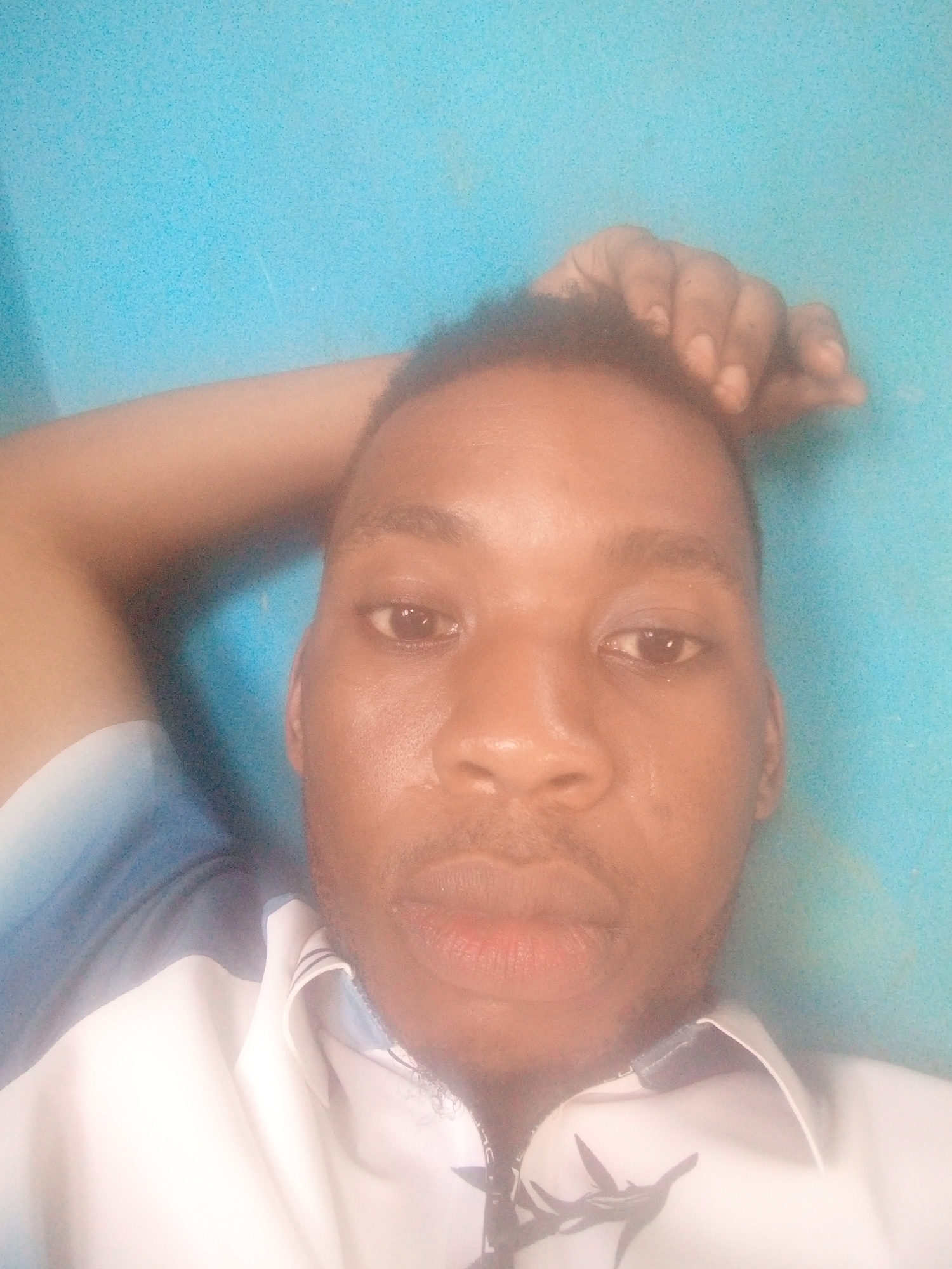 A man is posing for a selfie. He has his hand on his head and is wearing a white shirt with a blue background. The location is presumably Ikorodu, Nigeria.