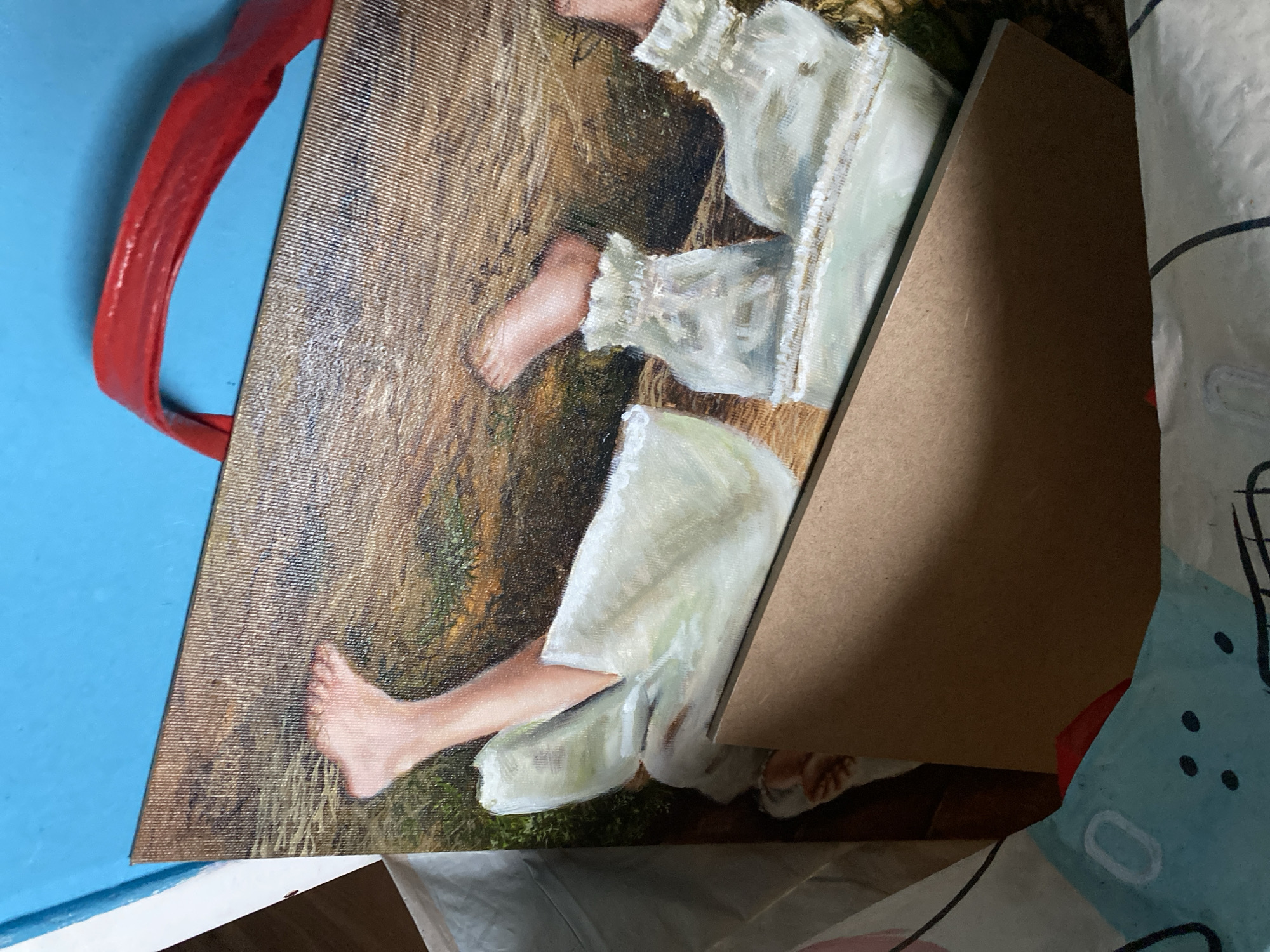 The image shows a close-up of a painting on canvas, with a red strap attached to the top. The painting depicts a close-up view of bare human feet and legs, clad in white, ruffled garments, lying on a textured, earthy background with hints of green. A piece of brown cardboard is propped against the painting. The painting is placed on top of a blue surface, and what appears to be a bag with a cartoonish print is visible at the bottom. The image's focus is on the painting and its immediate surroundings. There is no discernible information within the image to determine its location in the city of Bílina, Czechia.