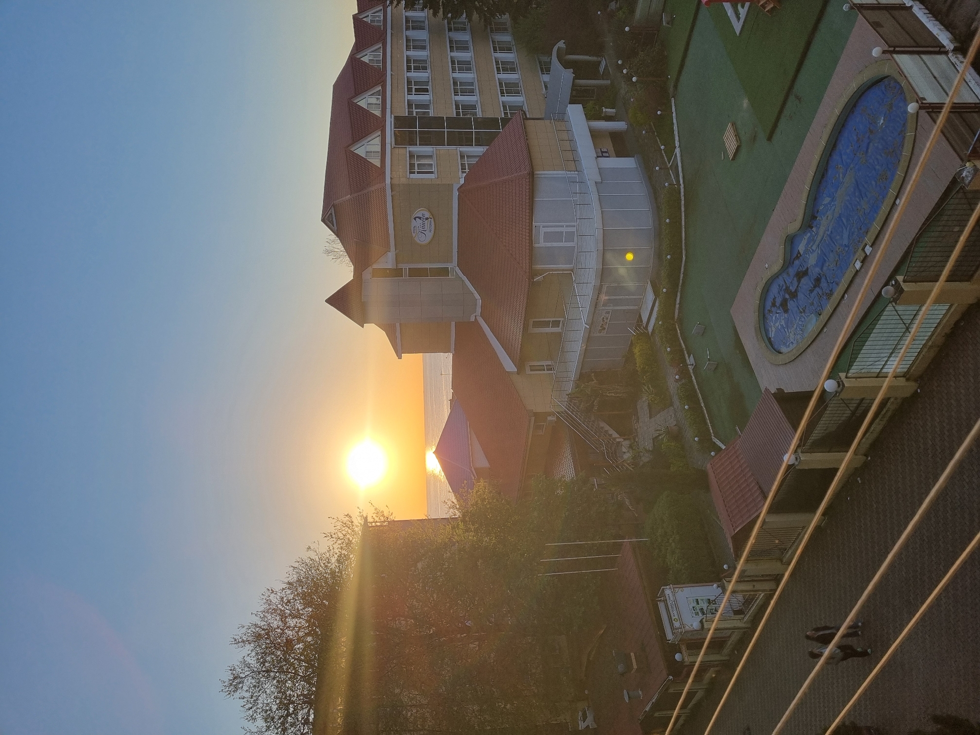 The image shows a view, possibly from a high vantage point, of a building complex with a hotel and pool area against a backdrop of a sunset over the sea. The primary building is a multi-story hotel with a red-tiled roof and light-colored facade. A pool with a blue mosaic design is visible, along with a surrounding landscaped area. In the foreground, there are power lines and glimpses of other buildings and greenery. The sunset creates a bright glow on the horizon. Based on these observations, it's difficult to determine the exact location in Sochi without additional information; however, the architectural style, presence of a hotel with a pool, and coastal location align with features of coastal resort areas.