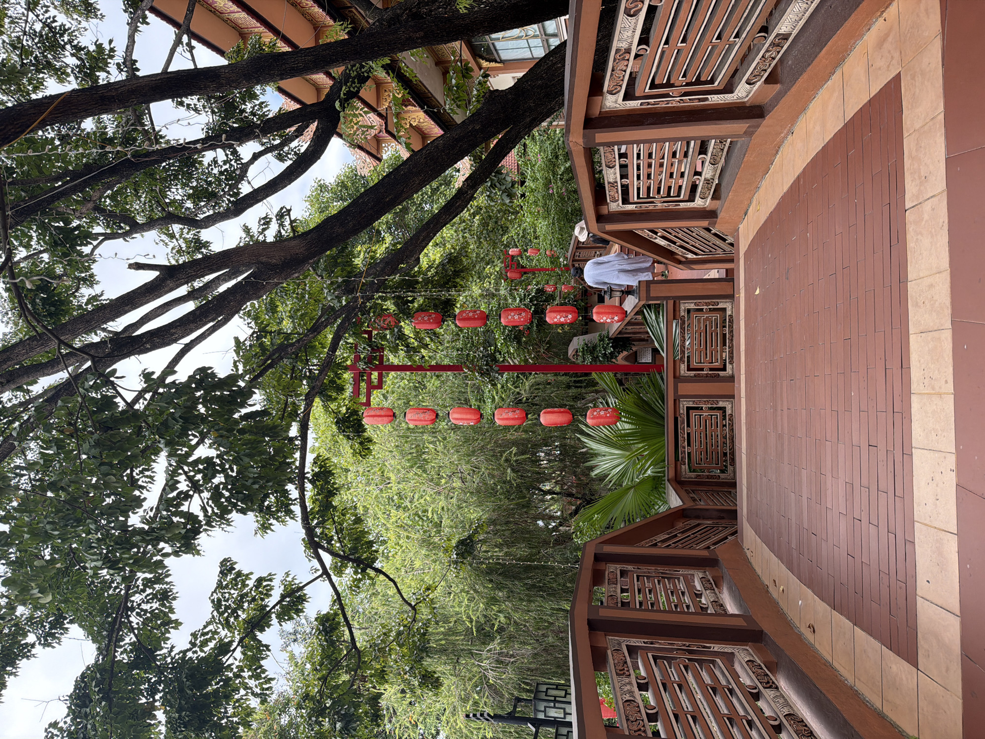 This image depicts a wooden bridge with intricate carvings on its railings, leading over a small body of water or decorative landscaping. The bridge is paved with reddish-brown tiles in a plank pattern, bordered by lighter tiles. Hanging from a red frame structure are several red paper lanterns with decorative patterns. To the right of the bridge, a building with a distinct orange and gold roofline and ornate architectural details is visible, characteristic of traditional Vietnamese or Chinese-inspired temple or pagoda architecture. Lush green trees and foliage surround the scene, contributing to a serene, possibly tranquil, atmosphere. One person, wearing a white garment, is seen walking away from the camera up the bridge, and another person, wearing a hat, is walking in the same direction further up the bridge. The scene is consistent with locations found in Hoi An, Vietnam, known for its well-preserved ancient town and cultural sites.
