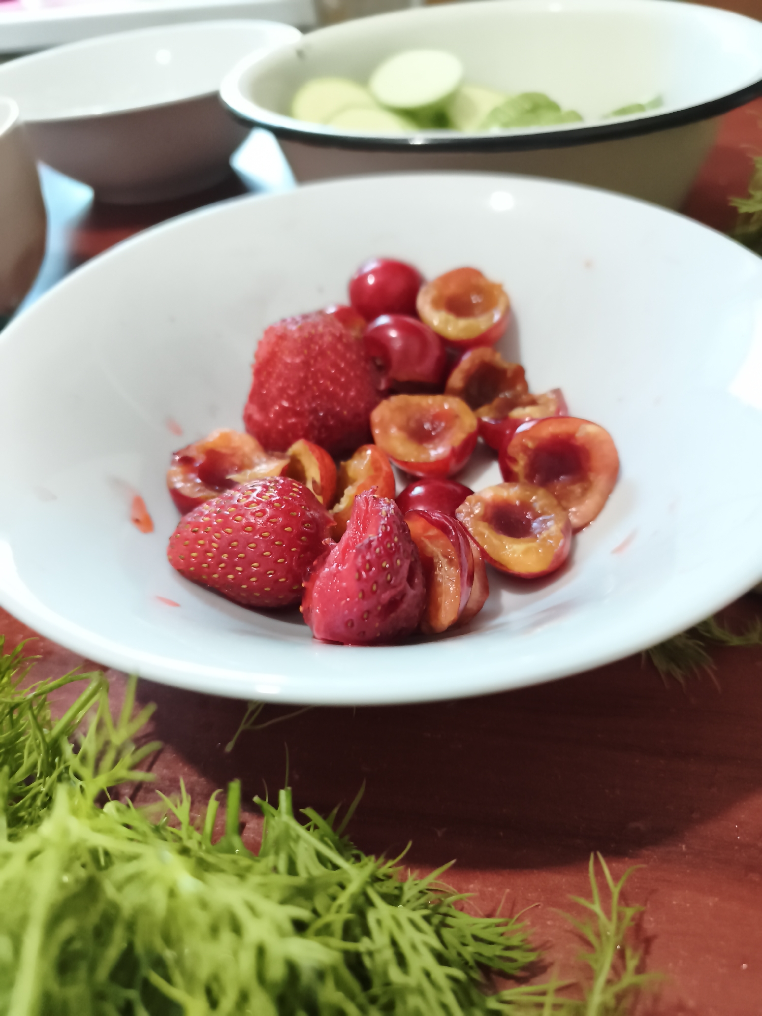 In Sokolivske, Ukraine, a white bowl sits on a dark red wooden surface, filled with sliced strawberries and cherries. The berries are fresh, some whole, some halved, revealing their bright red and orange interiors. Surrounding the bowl, a vibrant green dill garnish is scattered across the table. In the background, another white bowl contains sliced cucumbers. The lighting is soft, highlighting the textures and colors of the fruits.
