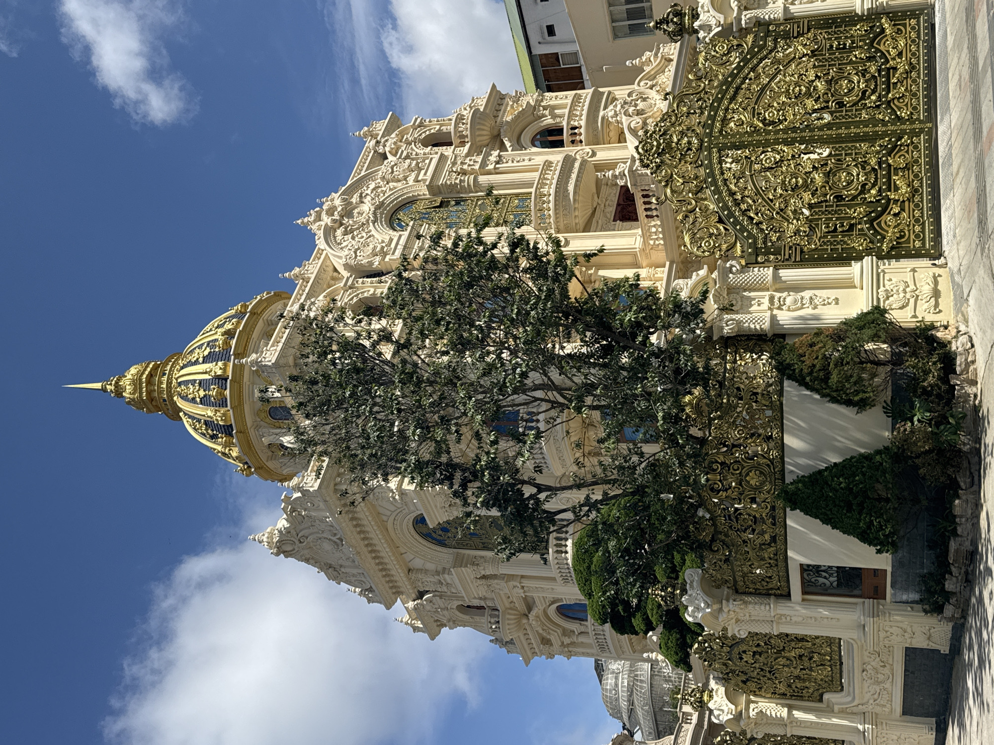 This is a full exterior shot of a large, ornate building in Da Lat, Vietnam. The building is cream-colored and features a prominent golden dome with blue accents, intricate architectural details, balconies, and large arched windows with gold ornamentation. A lush green tree stands in front of the building, partially obscuring the ornate golden gates that line the property. The sky is blue with scattered white clouds. The scene appears to be a private residence or a place of significant cultural or religious importance. The architectural style suggests a blend of European and Asian influences, possibly with a rococo or baroque inspiration.