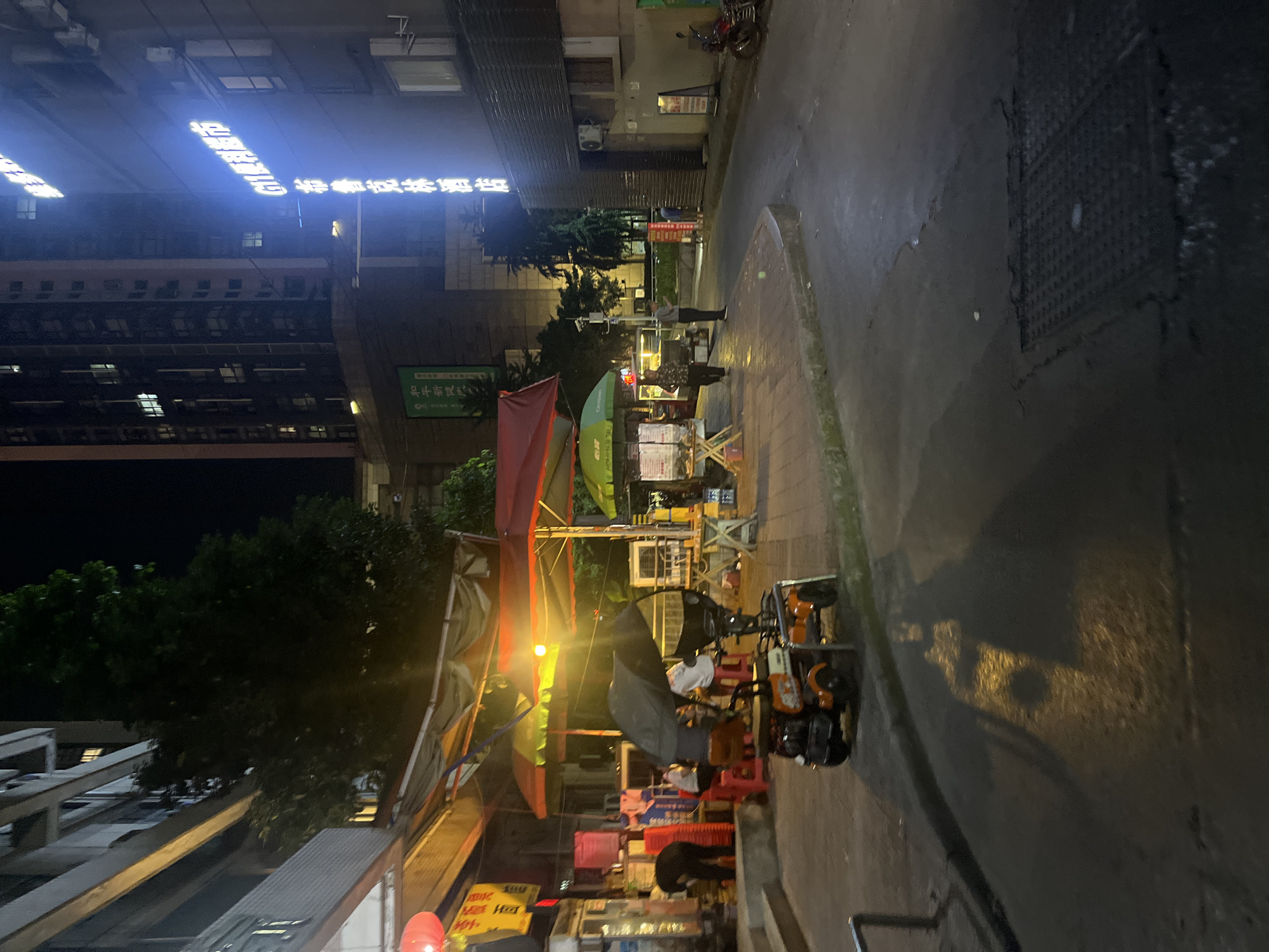This image depicts an illuminated urban street scene at night in Yuzhong District, China. The scene is dominated by a curved asphalt road with a sidewalk on the left.
On the left side, a multi-story building with visible balconies stands behind a series of temporary vendor stalls. These stalls are sheltered by various large umbrellas in colors including red, yellow, and green. Underneath these umbrellas, several individuals are present, some seated on red plastic stools and others standing. One person, dressed in dark clothing, is bent over near a container or bin on the far left. An orange three-wheeled utility vehicle, partially obscured by a black umbrella, is parked on the edge of the sidewalk or road in the mid-ground. Red lanterns are visible near the vendor area on the far left.
The right side of the image features a dark, multi-story building with prominent vertical neon signs in blue Chinese characters. The signs identify "G11 便利超市" (G11 Convenience Store) and "布鲁克林酒店" (Brooklyn Hotel). Further right, a motorcycle is parked on the street.
The road surface appears wet or reflective in several areas, showing light reflections, particularly in the foreground where a metal manhole cover is visible. Street lighting and illumination from the commercial establishments brightly light parts of the scene, creating strong contrasts with shadowed areas. In the background and along the left, dark foliage from trees is visible, bordering the urban structures. A small green sign with Chinese text "和平新城" (Heping New City) is partially visible on the left side, near some trees. Several other individuals are scattered throughout the scene, some appearing to be walking or standing near the stalls and along the road.