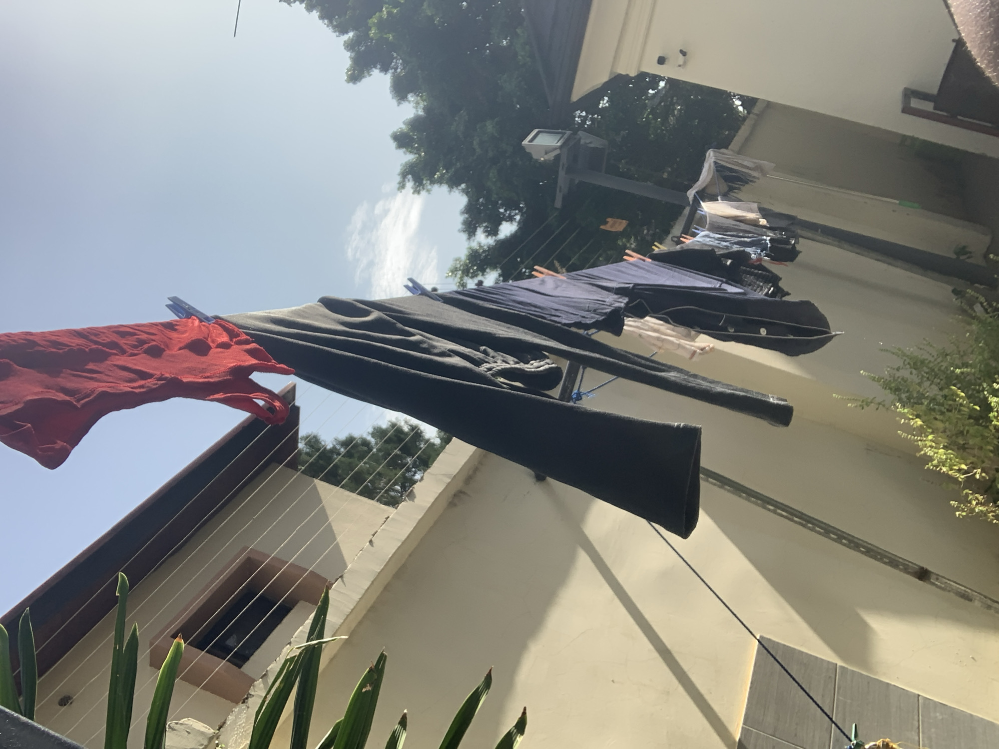 In an outdoor setting in Abuja, Nigeria, a clothesline is strung with various items of laundry. A bright red garment, possibly a shirt or scarf, is prominently displayed at the top, secured by a blue clothespin. Below it hang dark-colored garments, including a long-sleeved shirt or dress and a pair of trousers. Other items of clothing, in lighter colors and patterns, are also visible further down the line. The clothesline is attached to a light-colored wall with a brown trim and a window. Green foliage from a plant is visible in the foreground on the left side of the image. The background shows a clear sky with scattered clouds, lush green trees, and a lamppost with a small surveillance camera. The overall impression is a domestic scene of laundry drying in the sun.