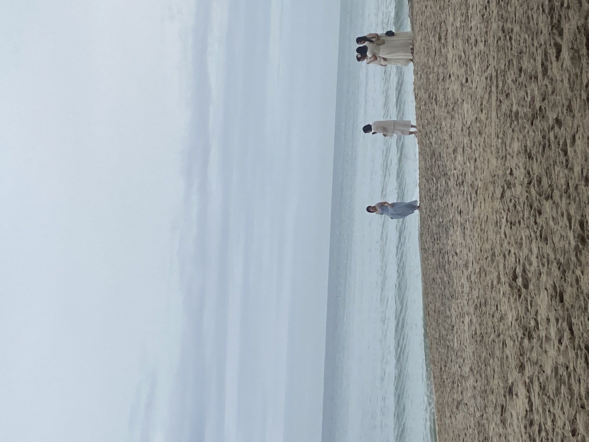 The image shows a group of people on a beach in Phu Thuy Ward, Vietnam. On the right, there is a large expanse of sandy beach. To the left is the ocean with a cloudy sky above. There are several people visible on the beach: a group of about four people wearing light-colored clothing are standing together, further down the beach there is another person in light colored clothing, and even further down is a person in a light blue dress. The people appear to be engaged in some kind of activity on the beach.