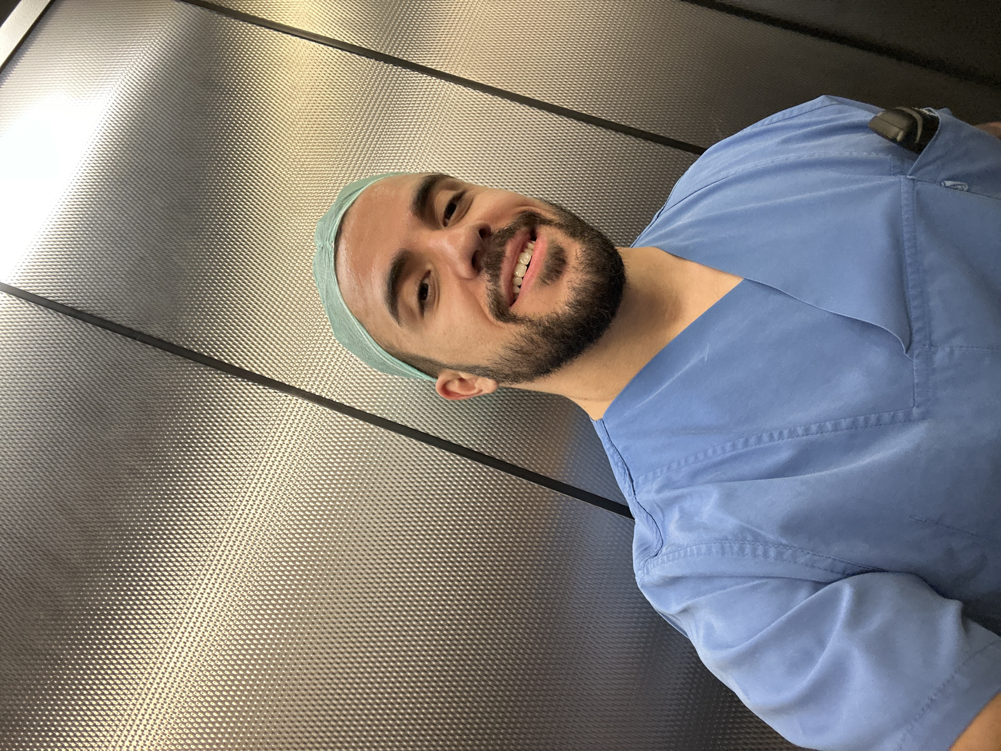 This media file is a portrait of a man wearing a light blue scrub suit and a light green surgical cap. He has dark hair, a beard, and is smiling broadly, showing his teeth. He appears to be in a casual setting, possibly a break room or a hallway, with a textured metallic wall in the background. The lighting suggests it is daytime. There is no visible text in the image. The man's relaxed posture and smile indicate a positive and friendly demeanor. The metallic wall has a repeating textured pattern, possibly indicating an industrial or clinical environment. The overall impression is of a health professional taking a moment for a self-portrait.