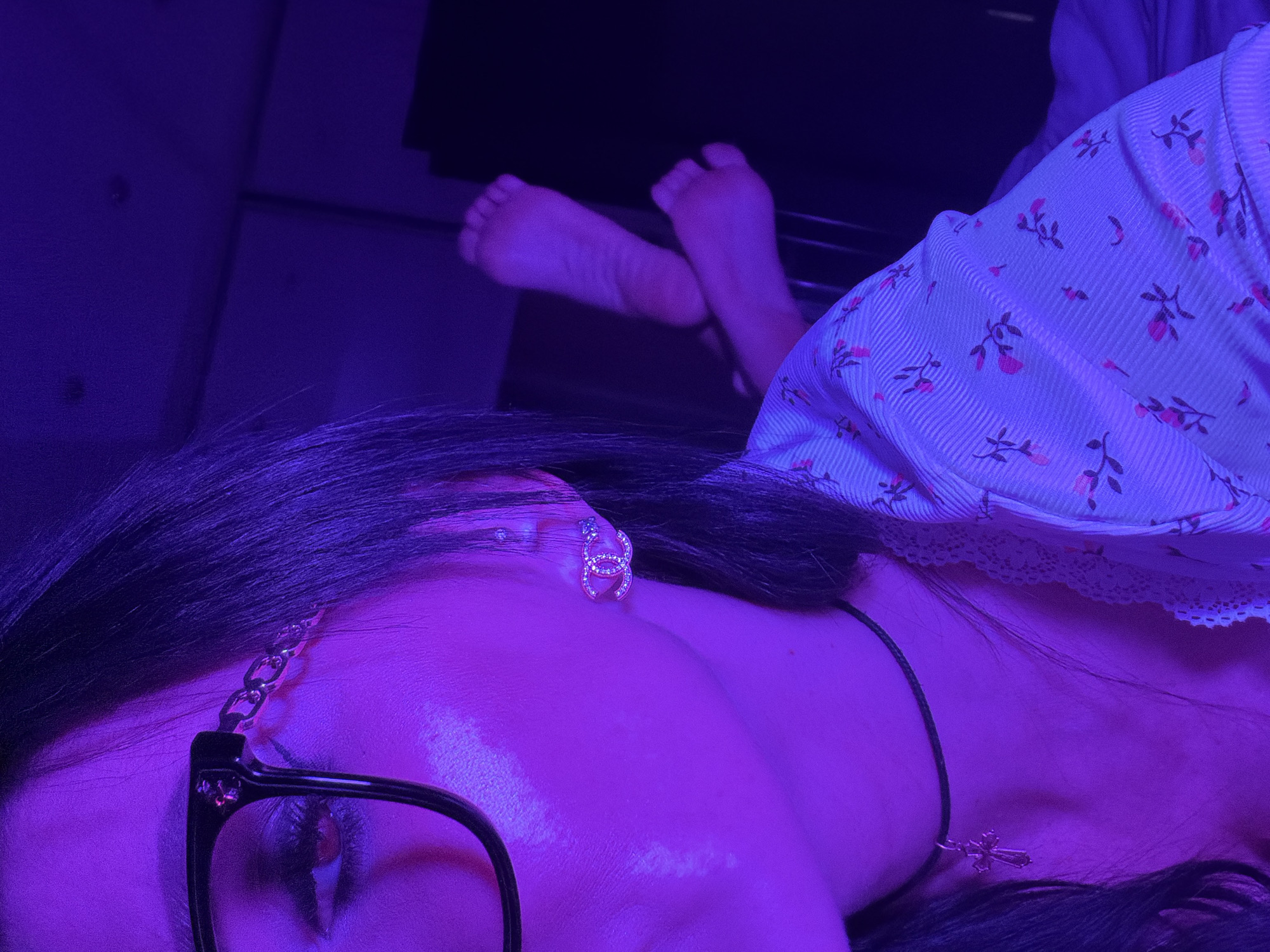 A close-up shot, bathed in purple light, depicts a person lying down. The person wears glasses, an earring resembling a logo, and a delicate necklace with a cross pendant. Their feet are visible in the background. The person is wearing a floral-patterned top. The location is not specified.