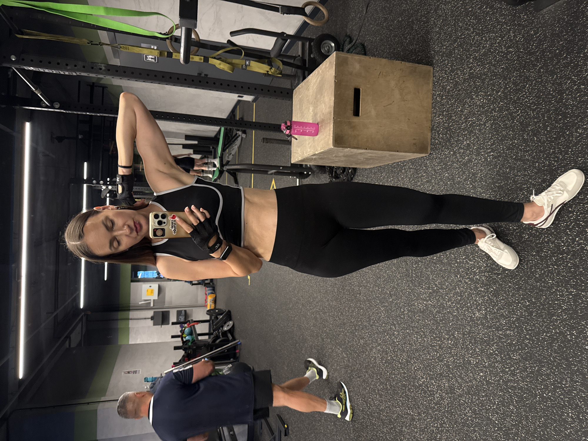 The image shows a woman in a gym, taking a selfie in a mirror. She is wearing a black sports bra and black leggings, with white sneakers. She has on black fingerless gloves. The gym appears to be well-equipped with various exercise machines and equipment, including a plyometric box, resistance bands, and weights. There is another person in the background, a man, also in gym attire. The lighting suggests it could be daytime, as there are bright overhead lights. The woman's expression is relaxed and she appears to be in good spirits. On her phone, there are several stickers, including one with a heart and flowers, and another that says "GOOD VIBES ONLY". The overall setting is a modern fitness facility.