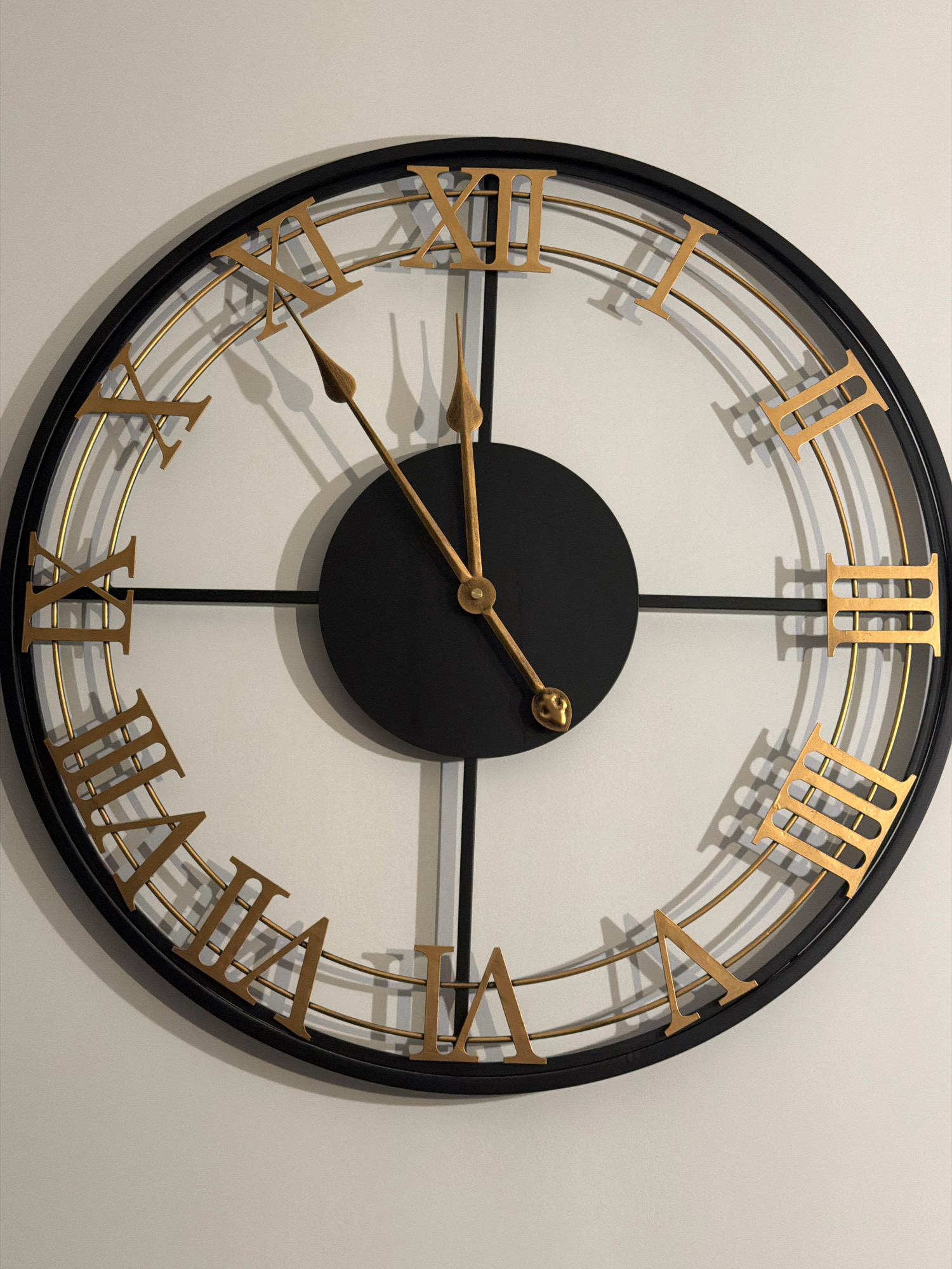 The image shows a close-up of a large, black-framed wall clock with a white face. The clock features gold-colored Roman numerals for the hour markers and gold-colored hour and minute hands. The hour hand is pointing towards the Roman numeral IX, and the minute hand is pointing towards the Roman numeral XII. The clock is hanging on a plain, light-colored wall, casting a subtle shadow. There are no people or any other objects visible in the image. The scene appears to be an indoor setting. The location in the city of Vniissok, Russia, is not discernible from the provided image.