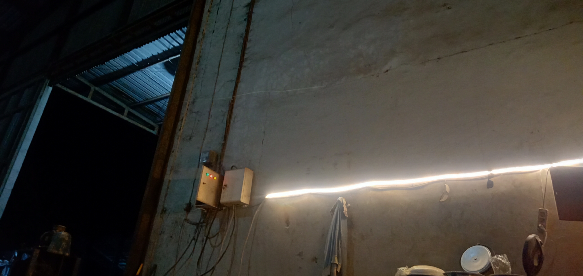 The image depicts an interior scene in a dimly lit structure in an unidentified city. The dominant features are a concrete wall and the corner of a building. Electrical wiring and two electrical boxes are affixed to the wall, one with lit indicator lights. An illuminated light strip runs horizontally along the wall, casting a glow on the surrounding area. Hanging on the wall is a cloth. Below the light strip, there are miscellaneous objects on a surface, including kitchenware. The left side of the image reveals a darker area with structural supports and a metal roof.
