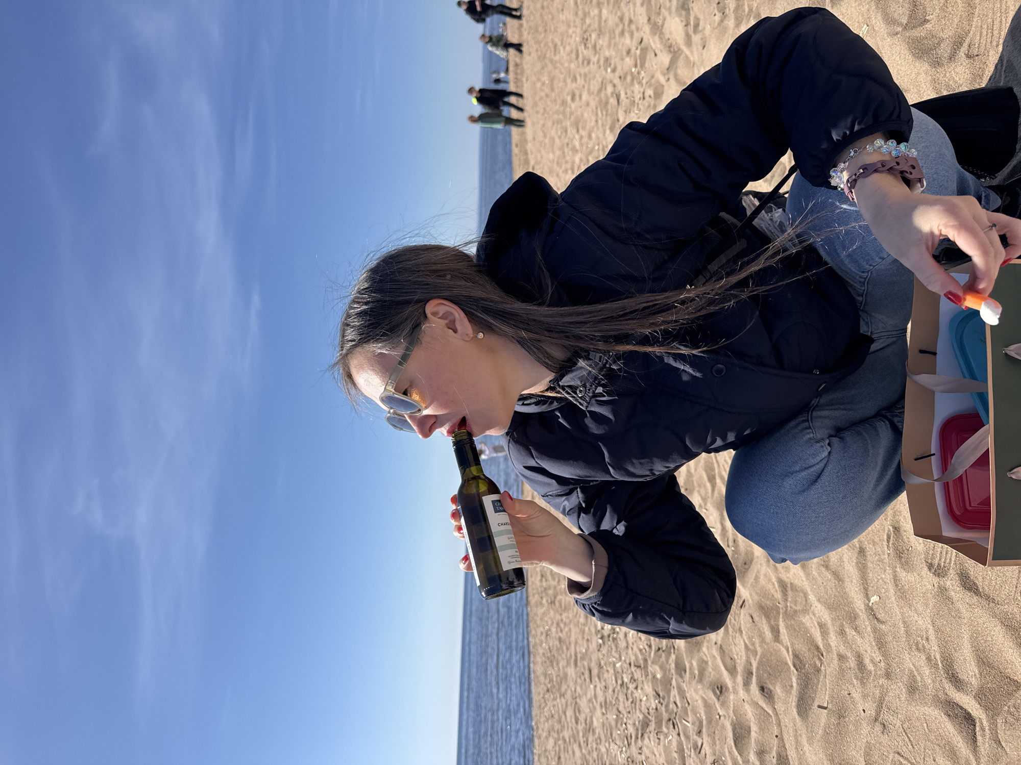 A woman is squatting on a sandy beach next to water, possibly the Gulf of Finland near Komarovo, Russia. She is wearing a dark jacket, jeans, and sunglasses. She has long brown hair and is holding a bottle of wine to her lips. In her other hand, she holds a small orange and white object over a bag that appears to contain food or other items. In the background, some people are walking on the beach. The sky is blue with a few wispy clouds.