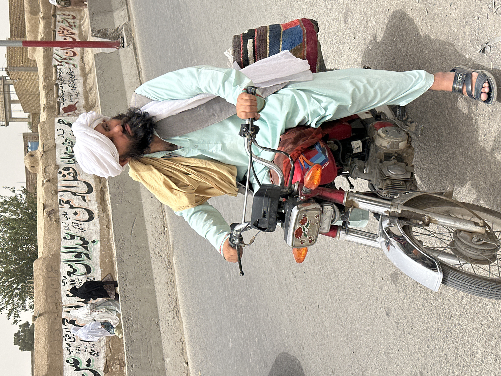 In Kuchlak, Pakistan, a man with a dark beard and a white turban rides a red motorcycle. He wears a light blue shalwar kameez and a grey vest, with a tan scarf draped over his shoulders. On the back of the motorcycle, a colorful woven bag is attached. The man is on a paved road, with a concrete barrier to his left. Behind the barrier, a wall displays Arabic calligraphy and images of people. The background shows buildings and trees under a bright sky.