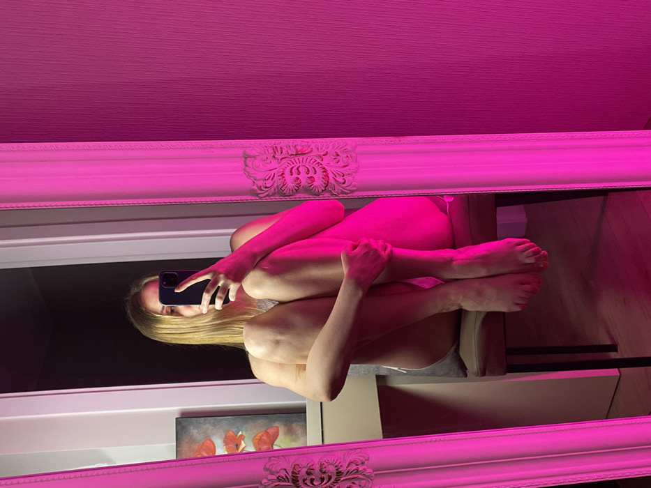 A mirror reflects a woman sitting cross-legged, knees drawn up, holding a phone. The woman's blonde hair is visible, and her face is obscured by the phone. A decorative frame surrounds the mirror, and pink light illuminates the scene. A window and a painting with red flowers can be seen in the background, along with light-colored furniture.